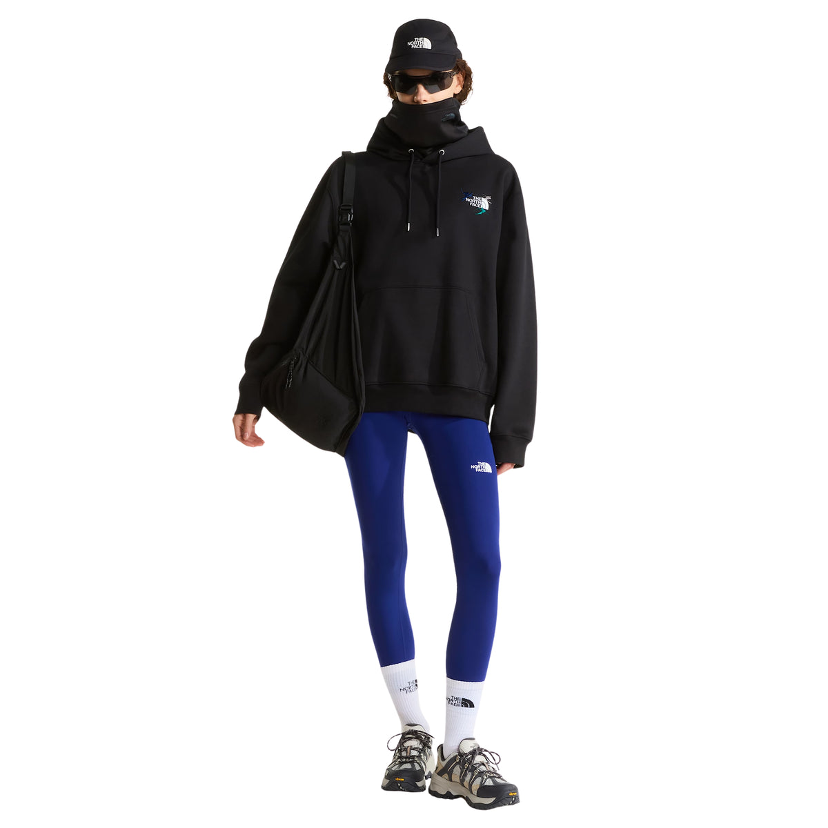 Modelo de corpo inteiro com sweat The North Face Off Piste Embro preta, leggings azuis e bolsa preta ao ombro.
