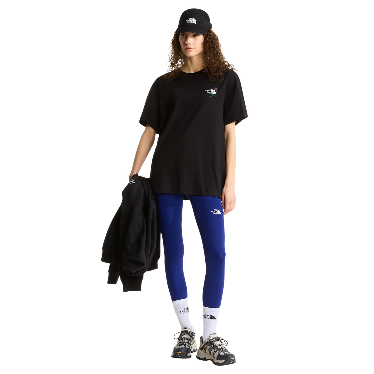 Modelo de corpo inteiro com t-shirt The North Face Off Piste Embro preta, leggings azuis e boné preto.