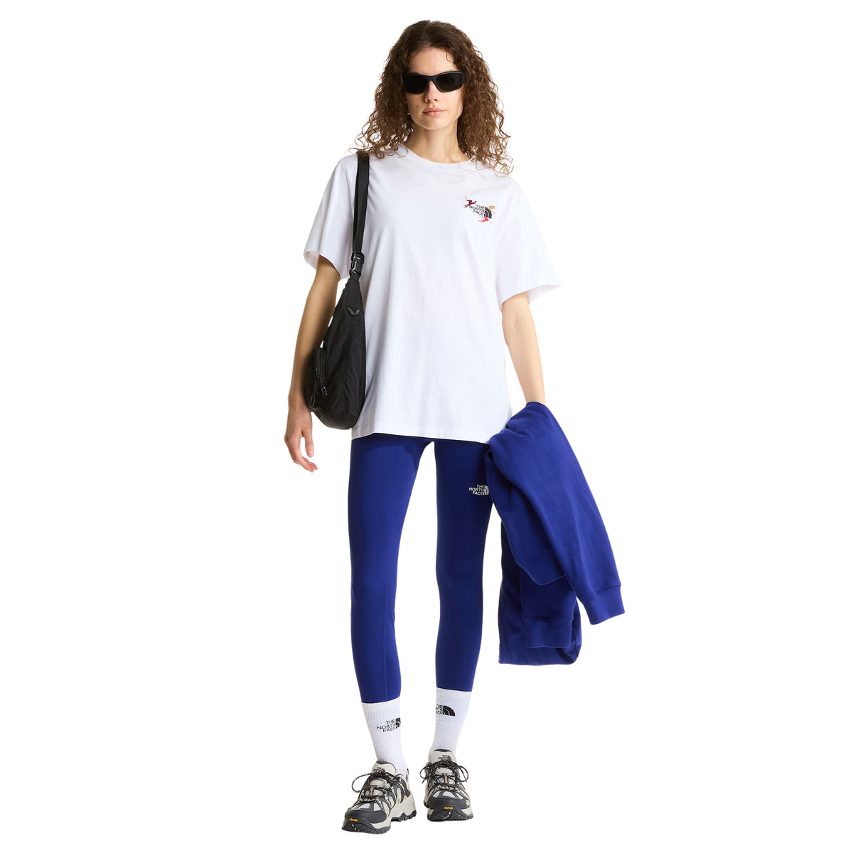 Modelo de corpo inteiro com t-shirt The North Face Off Piste Embro branca, leggings azuis e bolsa preta ao ombro.