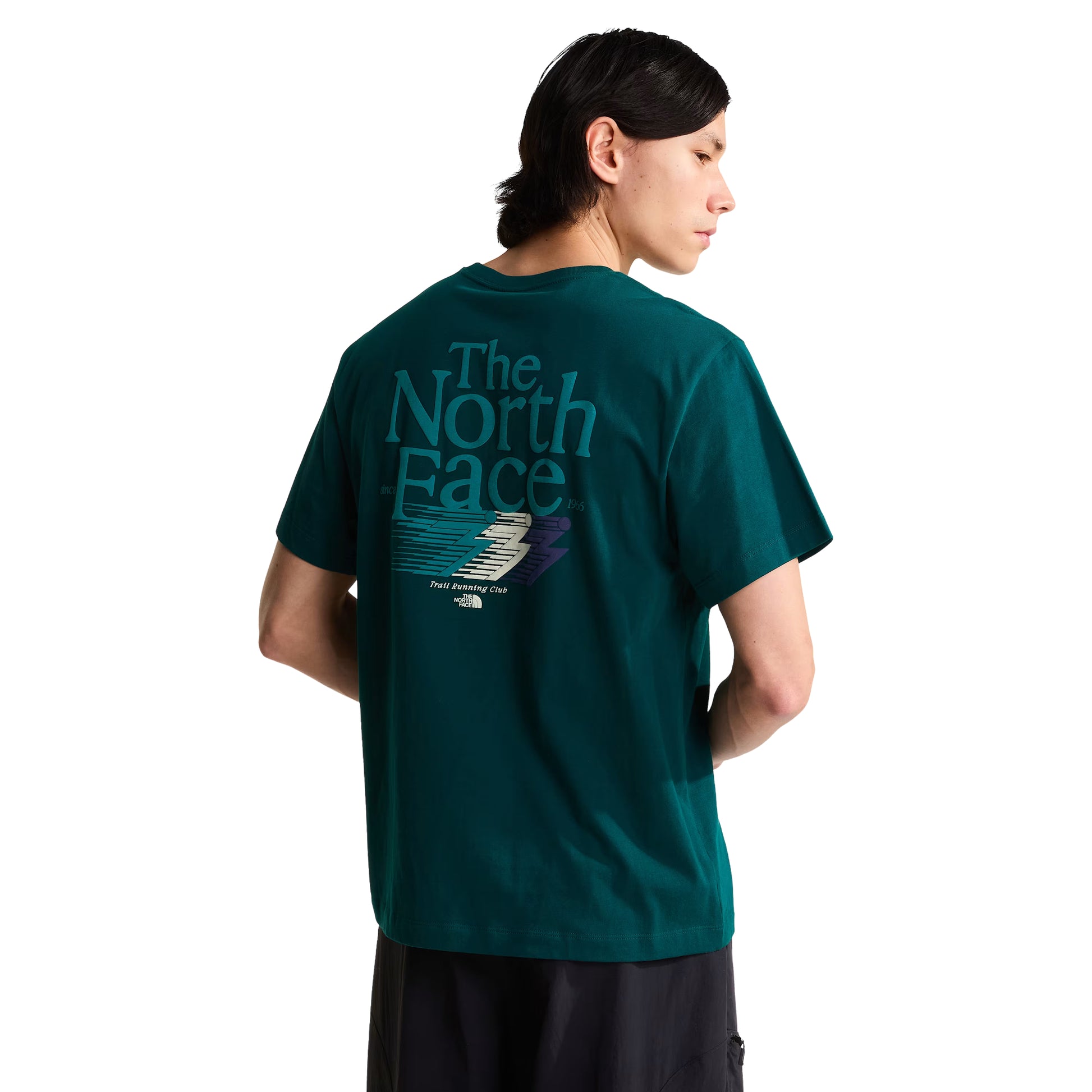 T-Shirt The North Face Retro Trail Relaxed em Pacific Teal — modelo masculino de costas, cabelo comprido escuro apanhado, calças pretas, gráfico "The North Face" em teal/azul-petróleo com inscrição "Trail Running Club" e raio estilizado em creme, teal e roxo nas costas visíveis, fundo branco.