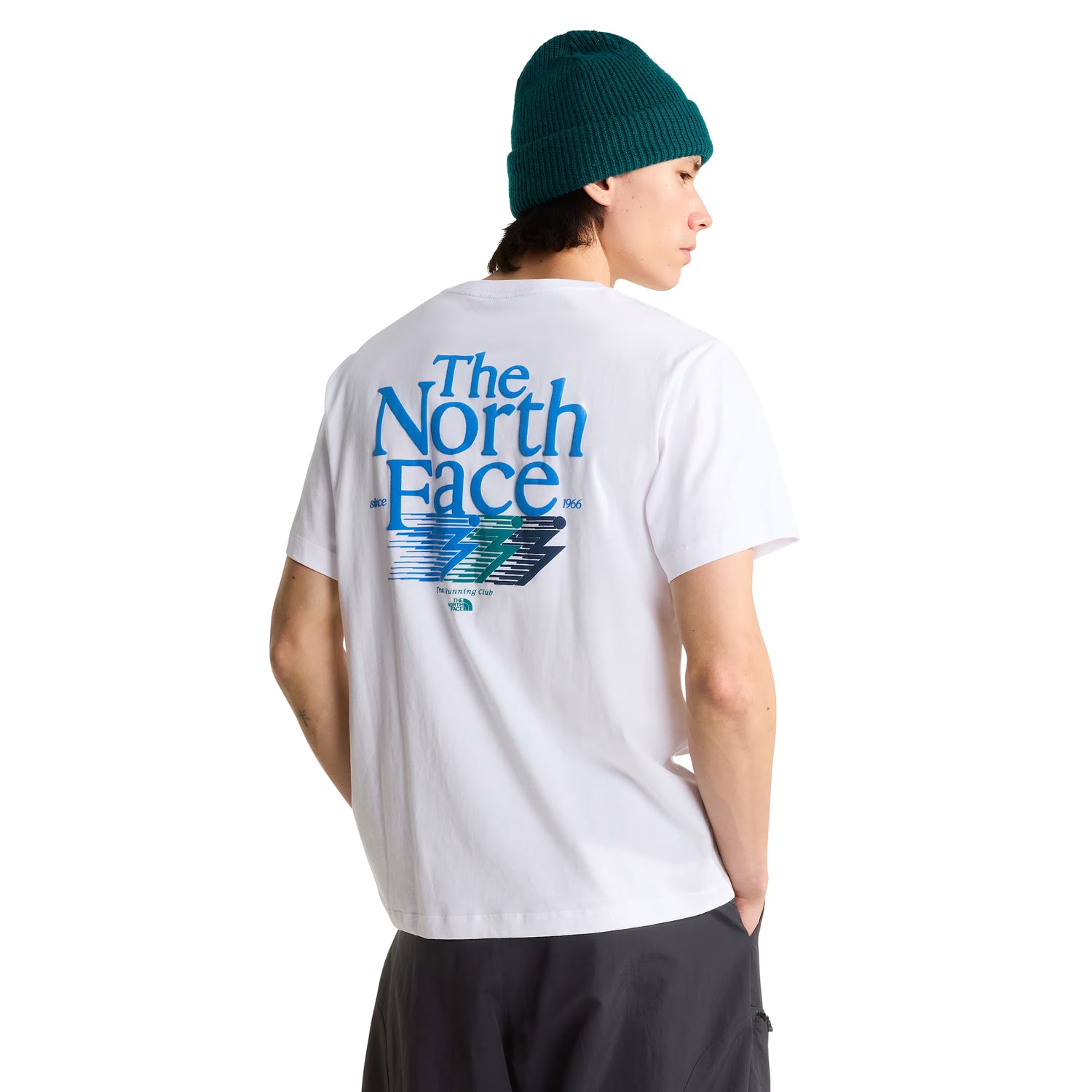 T-Shirt The North Face Retro Trail Relaxed em TNF White — modelo masculino de costas a 3/4, gorro TNF verde-teal, calças pretas, gráfico "The North Face" em azul-céu com raio estilizado em azul, verde e cinza-escuro e inscrição "Trail Running Club" nas costas visíveis, fundo branco.