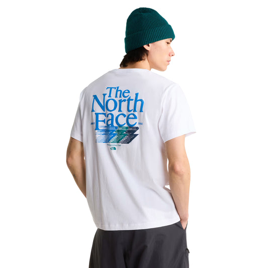 T-Shirt The North Face Retro Trail Relaxed em TNF White — modelo masculino de costas a 3/4, gorro TNF verde-teal, calças pretas, gráfico "The North Face" em azul-céu com raio estilizado em azul, verde e cinza-escuro e inscrição "Trail Running Club" nas costas visíveis, fundo branco.