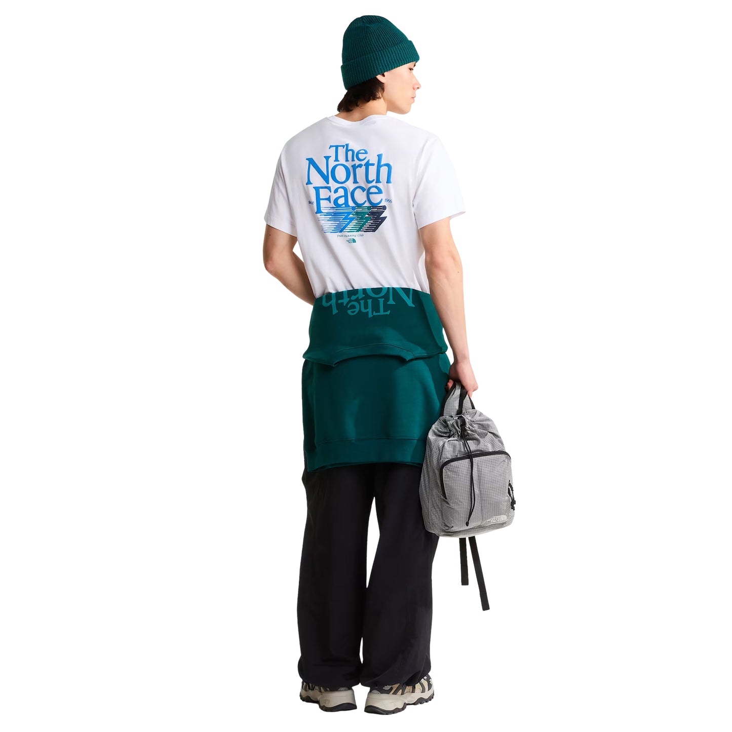 T-Shirt The North Face Retro Trail Relaxed em TNF White — modelo masculino de costas, corpo inteiro, gorro TNF verde-teal, sweatshirt TNF verde-teal atada à cintura, calças pretas largas e sapatilhas TNF bege, mochila cinza TNF na mão direita, gráfico Retro Trail nas costas visível, fundo branco.