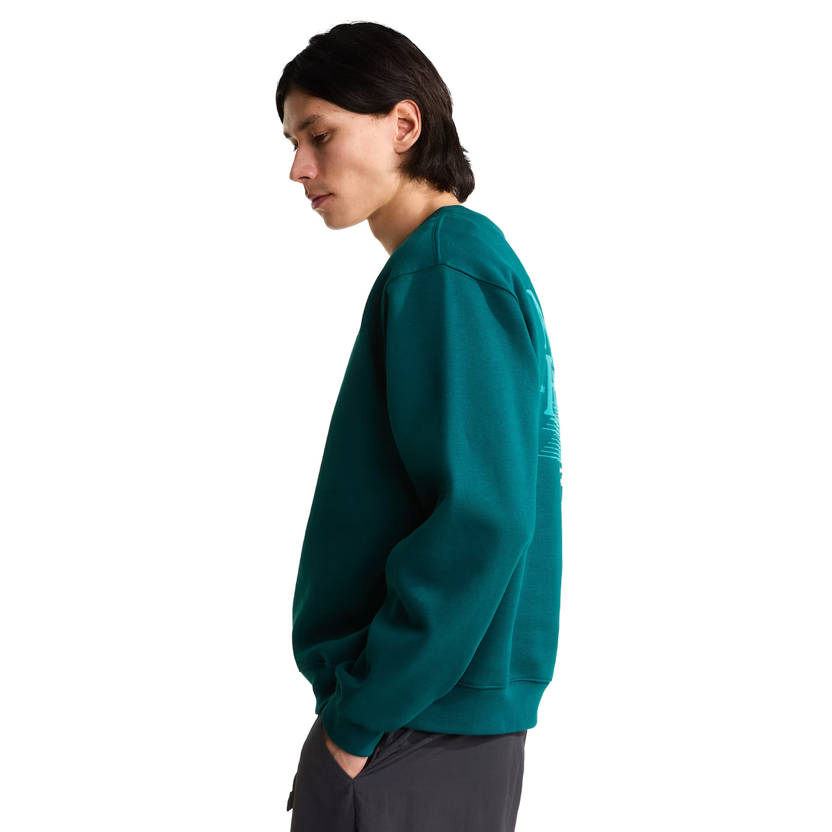 Sweatshirt para homem The North Face Retro Trail Relaxed Crew em Pacific Teal — modelo masculino de perfil esquerdo, corpo inteiro, malha polar teal, gráfico Retro Trail em relevo de camurça parcialmente visível nas costas, gola canelada, ombros descaídos, corte relaxed, calças cinzento escuro, mão no bolso, fundo branco.