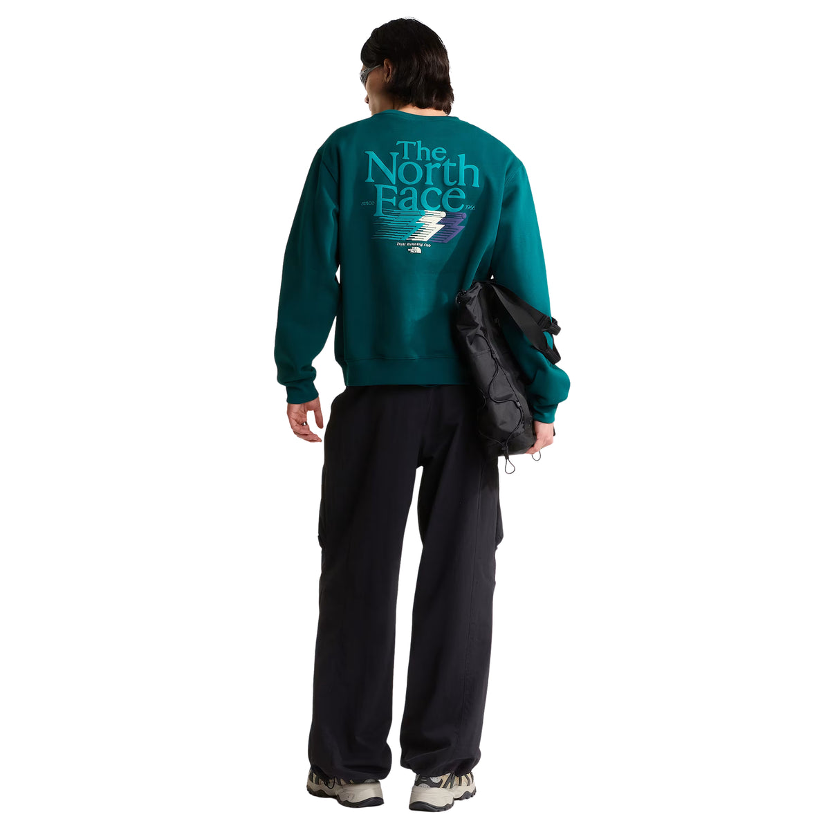 Sweatshirt para homem The North Face Retro Trail Relaxed Crew em Pacific Teal — modelo masculino de costas, corpo inteiro, malha polar teal, gráfico Retro Trail em relevo de camurça nas costas com "The North Face" em tipografia vintage e raio colorido, saco preto The North Face pendurado no ombro esquerdo, calças cargo pretas largas, sapatilhas de trail, fundo branco.