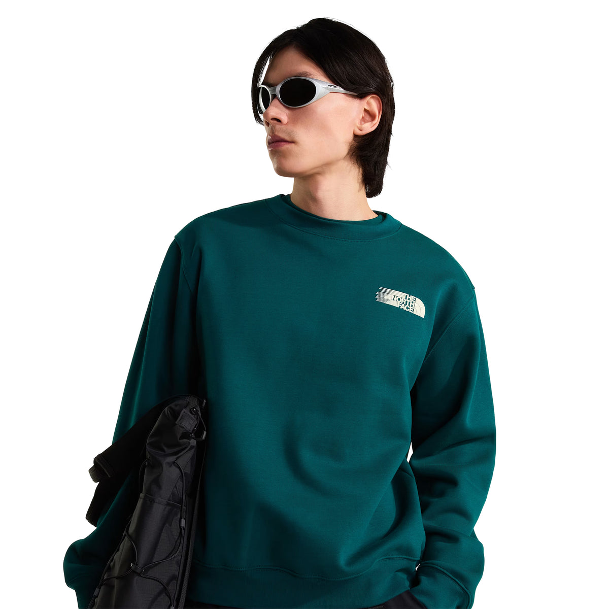 Sweatshirt para homem The North Face Retro Trail Relaxed Crew em Pacific Teal — modelo masculino de frente, meio corpo, malha polar teal, logo The North Face em relevo creme no peito esquerdo, óculos de sol ovais prata, saco preto The North Face pendurado no ombro, gola canelada, corte relaxed, fundo branco.