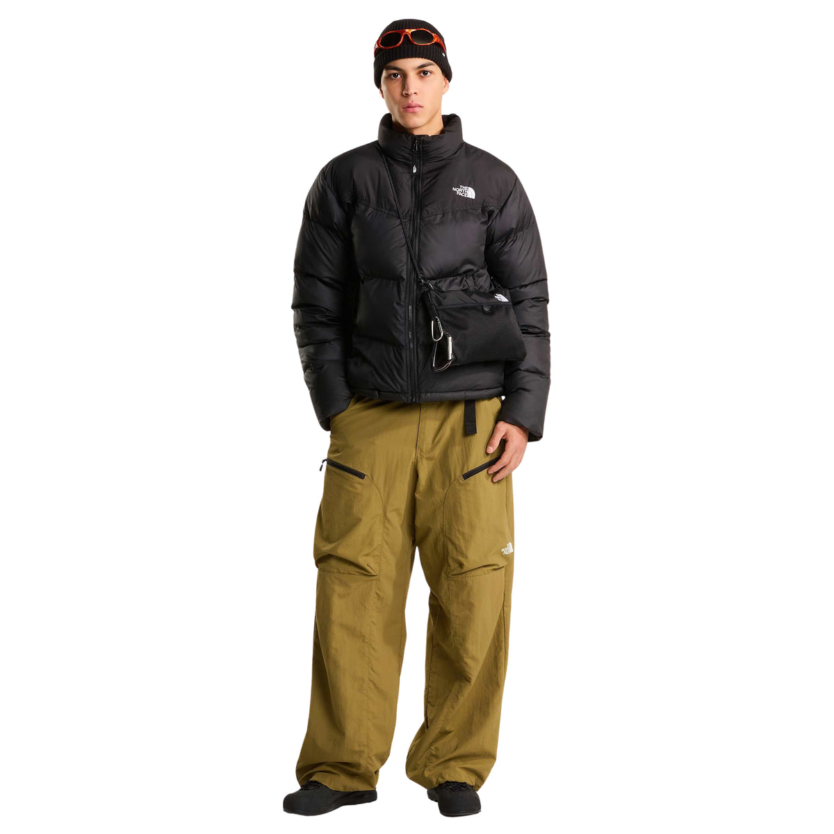 Modelo de corpo inteiro com casaco The North Face Saikuru preto, calças verde-azeitona, gorro preto e bolsa preta a tiracolo.
