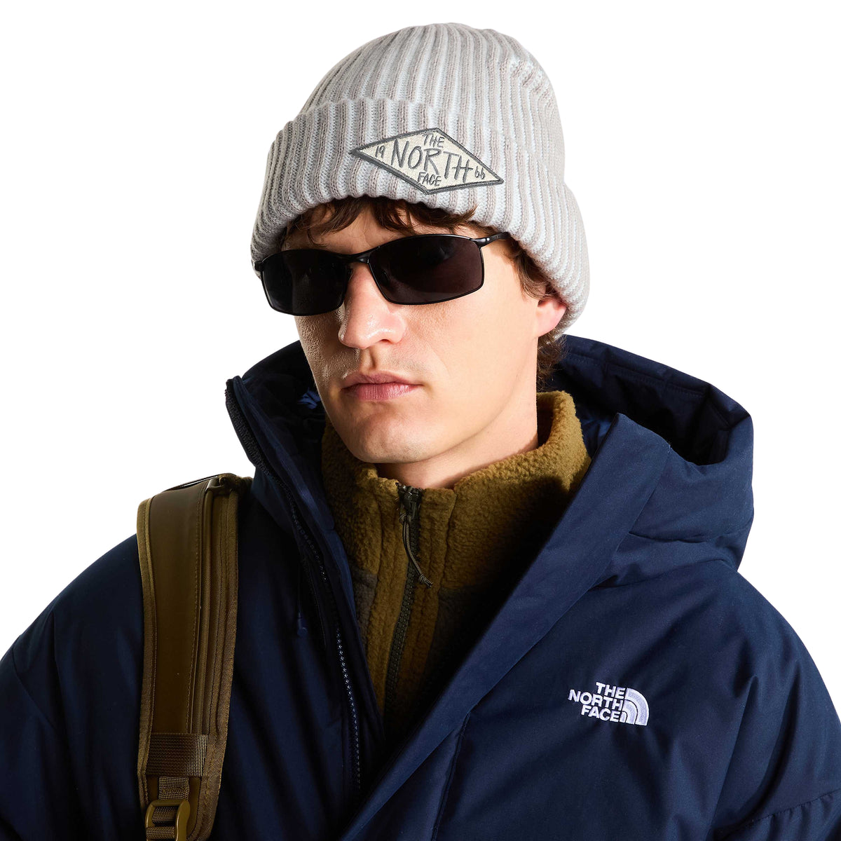 Modelo de frente com gorro The North Face Salty Lined SE Stone Slab, óculos de sol e casaco azul-escuro, com emblema TNF 66 visível.