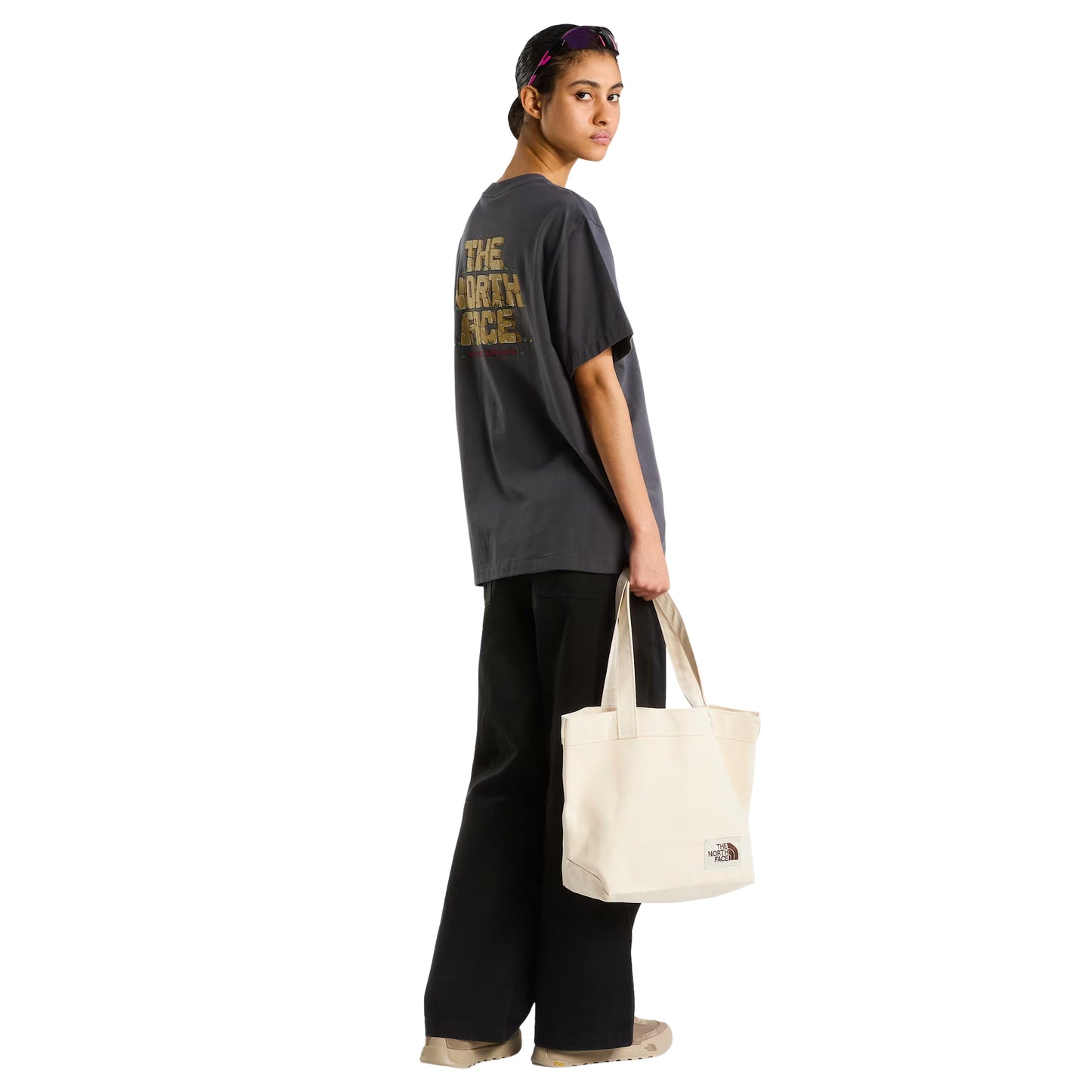 T-Shirt The North Face Stone Logo Relaxed em Anthracite Grey — modelo feminino de costas a 3/4, corpo inteiro, calças pretas largas e sapatilhas bege TNF, tote bag TNF em cru na mão direita, gráfico Stone Logo nas costas parcialmente visível, fundo branco.