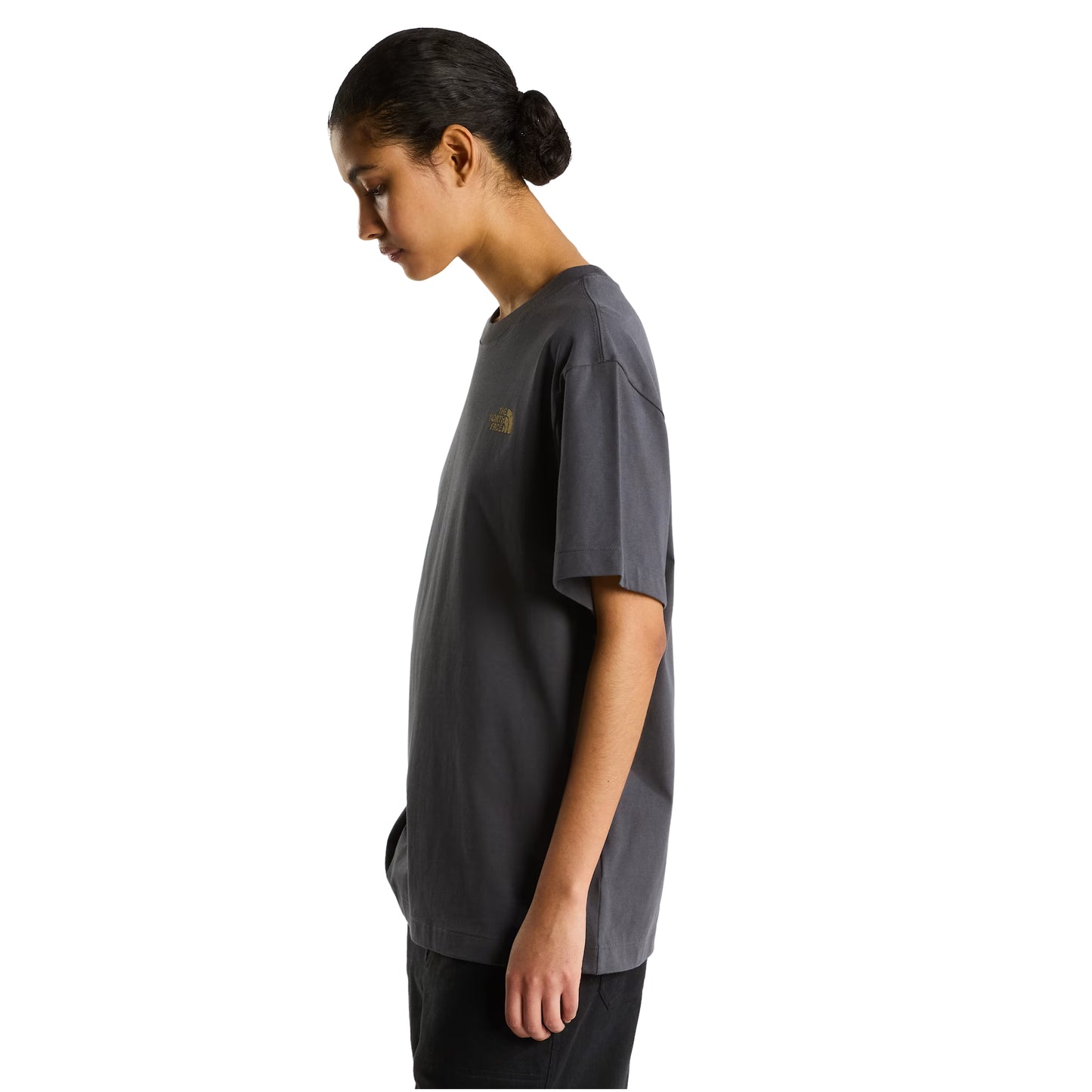 T-Shirt The North Face Stone Logo Relaxed em Anthracite Grey — modelo feminino de perfil esquerdo, cabelo apanhado, calças pretas, logo TNF em dourado parcialmente visível no peito, corte relaxed oversized, fundo branco.