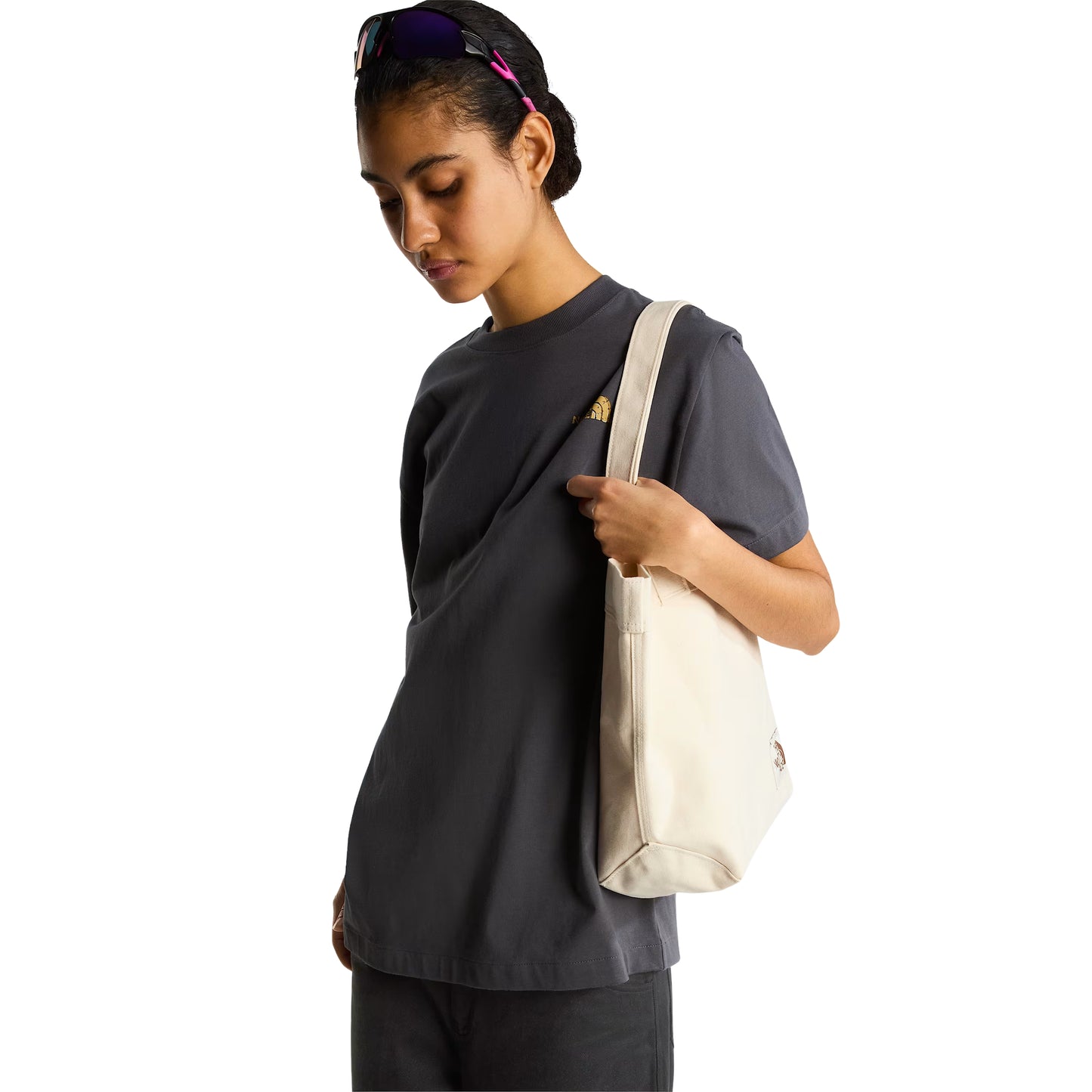 T-Shirt The North Face Stone Logo Relaxed em Anthracite Grey — modelo feminino de frente a 3/4, corpo inteiro, calças pretas largas, tote bag TNF em cru ao ombro esquerdo, logo TNF Stone em dourado no peito esquerdo visível, fundo branco.