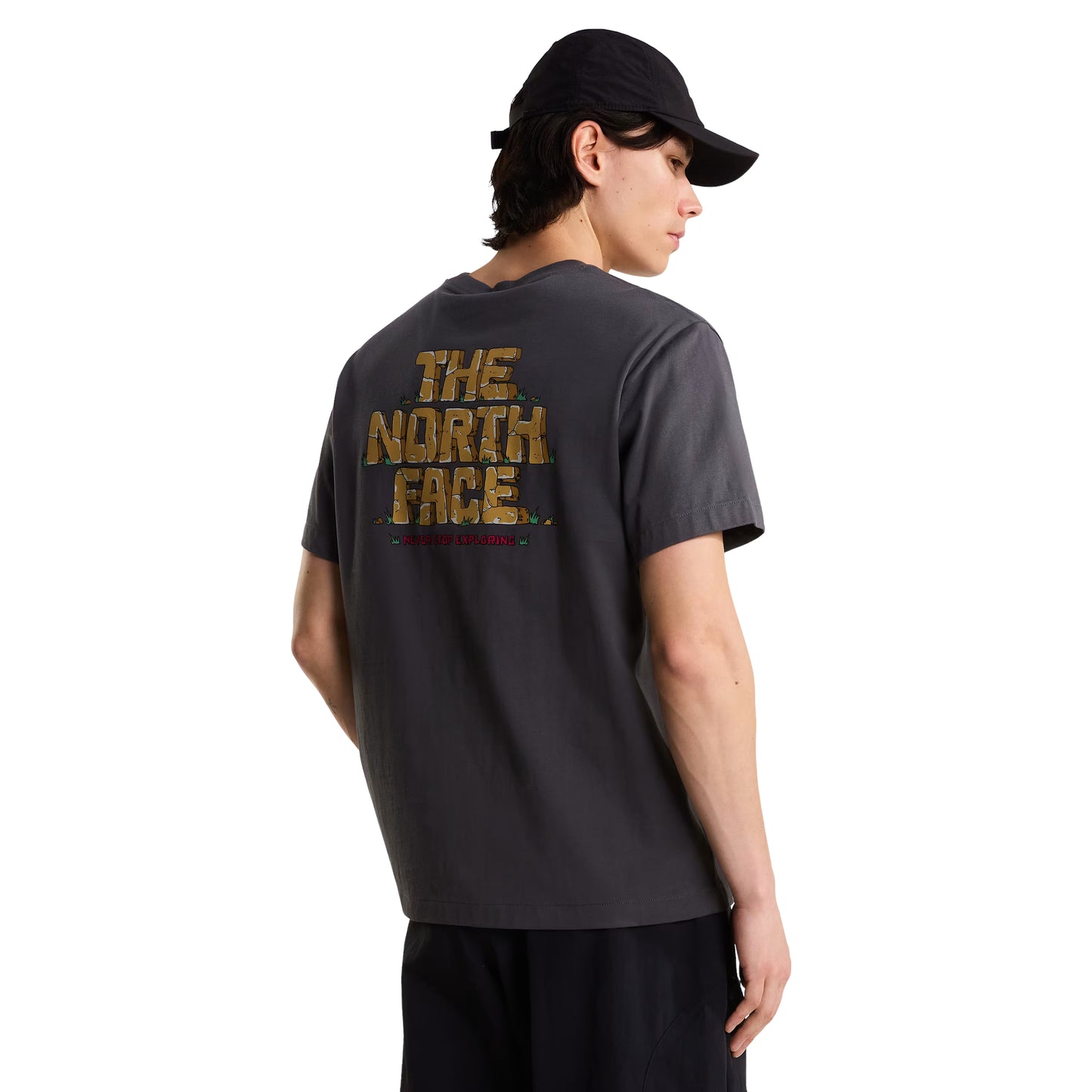T-Shirt The North Face Stone Logo Relaxed em Anthracite Grey — modelo masculino de costas, boné TNF preto, calças pretas, gráfico "The North Face" estilo pedra em dourado/ocre e inscrição "Never Stop Exploring" em vermelho nas costas visíveis, fundo branco.