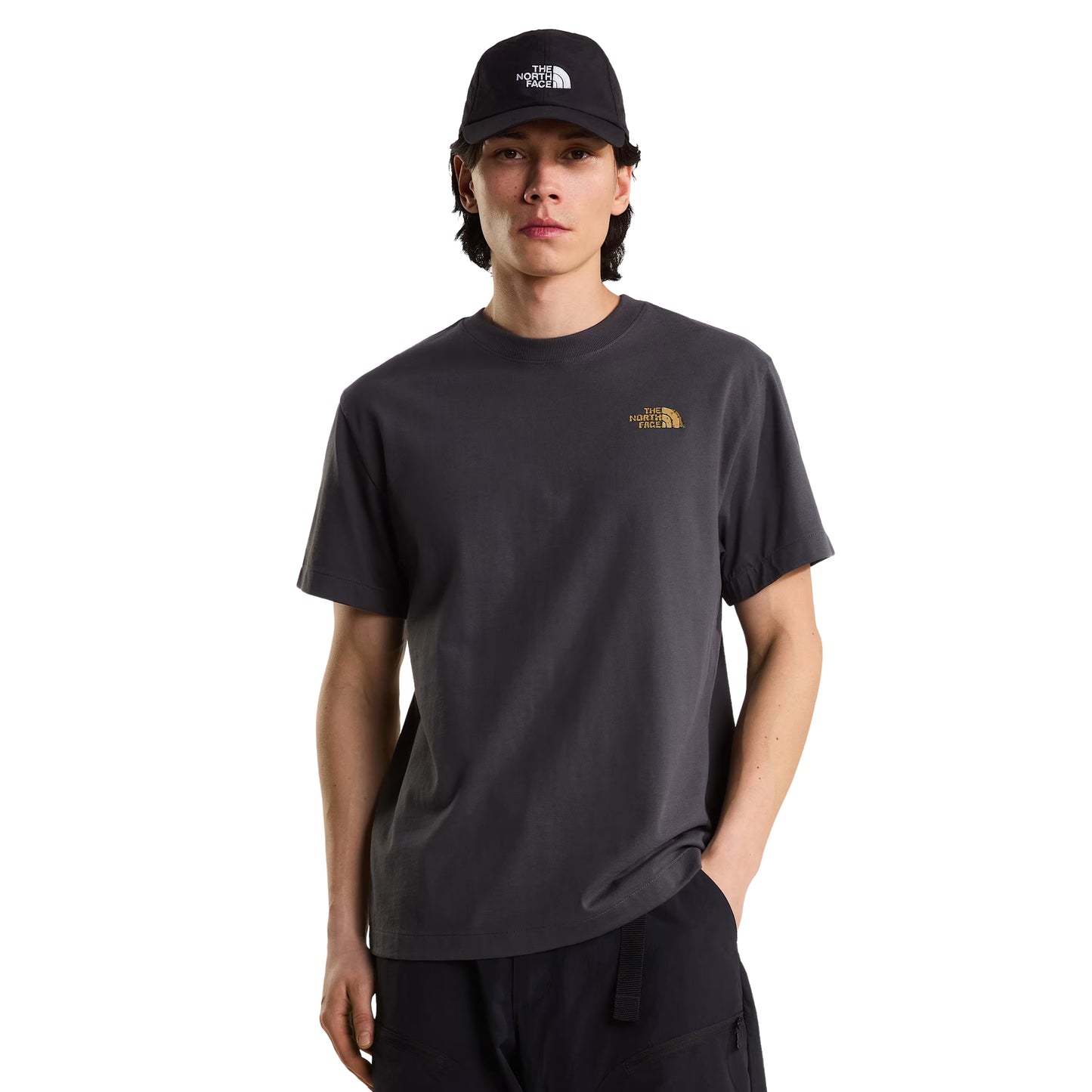 T-Shirt The North Face Stone Logo Relaxed em Anthracite Grey — modelo masculino de frente, boné TNF preto, calças pretas, logo TNF em dourado no peito esquerdo visível, corte relaxed, fundo branco.