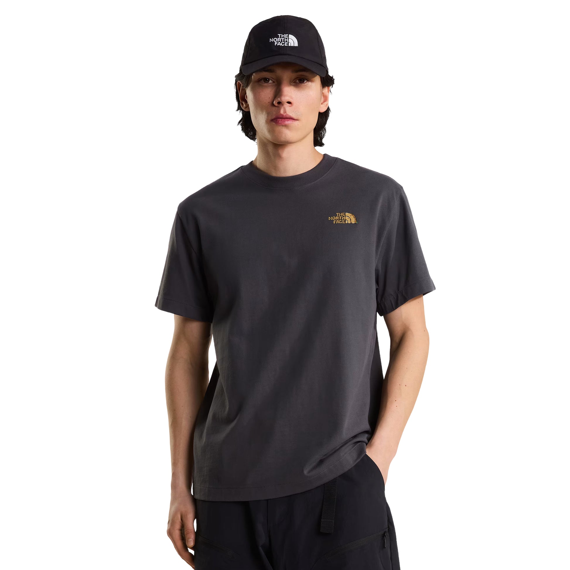 T-Shirt The North Face Stone Logo Relaxed em Anthracite Grey — modelo masculino de frente, boné TNF preto, calças pretas, logo TNF em dourado no peito esquerdo visível, corte relaxed, fundo branco.