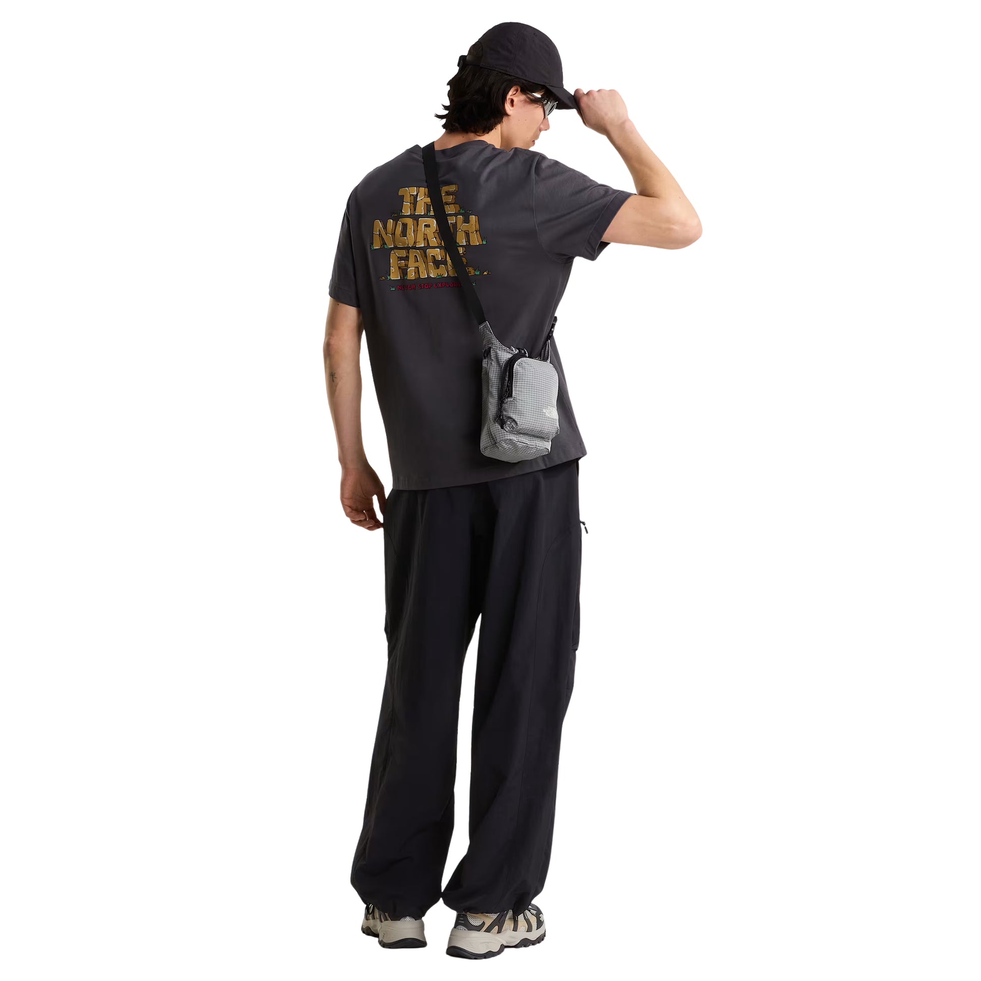 T-Shirt The North Face Stone Logo Relaxed em Anthracite Grey — detalhe das costas com modelo masculino, gráfico "The North Face" estilo pedra em dourado/ocre com detalhes verdes e inscrição "Never Stop Exploring" em vermelho na base, fundo branco.