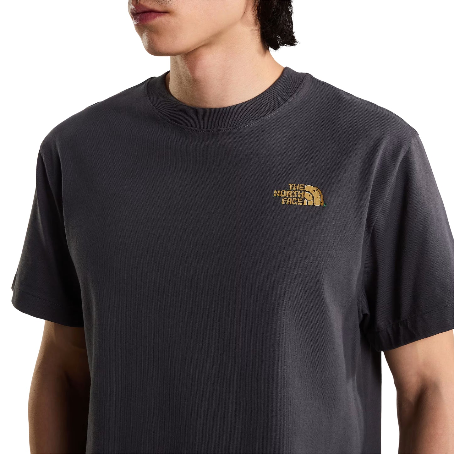 T-Shirt The North Face Stone Logo Relaxed em Anthracite Grey — detalhe das costas com modelo masculino, boné preto e alça de saco crossbody preta a cruzar as costas, gráfico "The North Face" estilo pedra em dourado/ocre e inscrição "Never Stop Exploring" em vermelho visíveis, fundo branco.