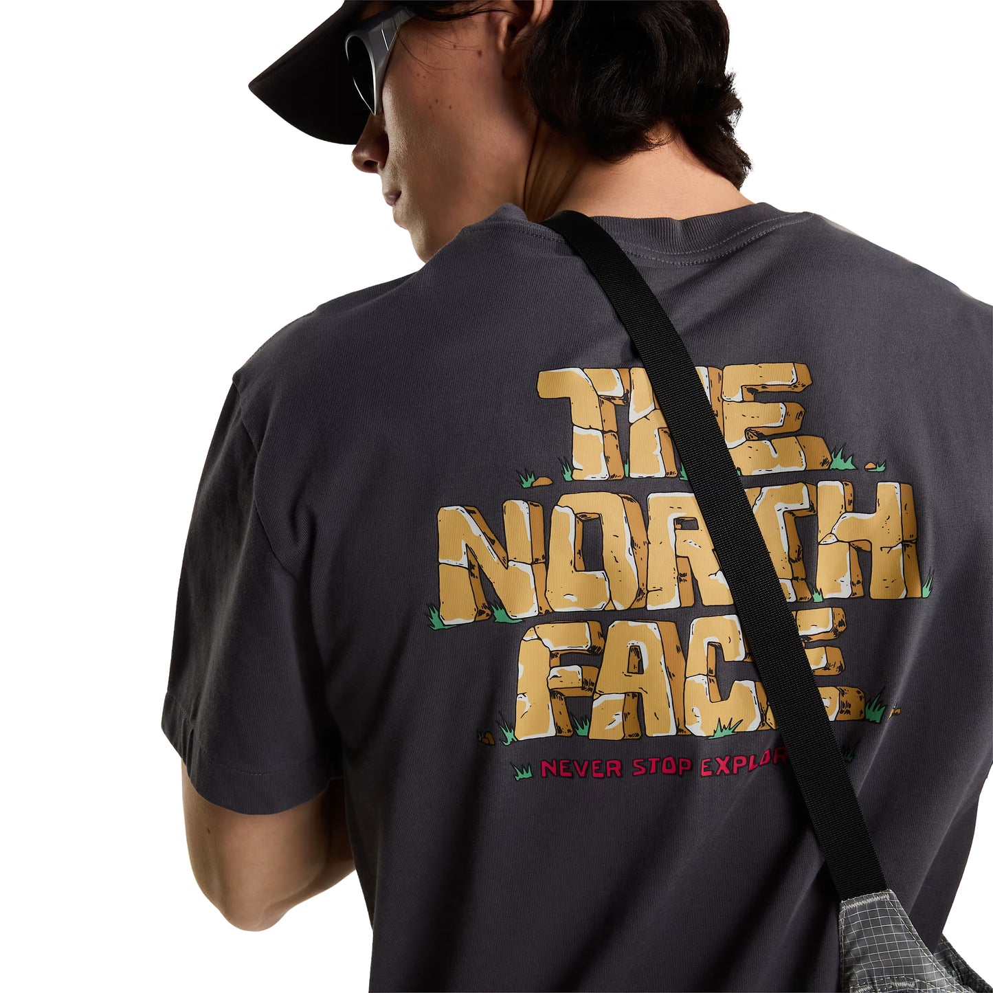 T-Shirt The North Face Stone Logo Relaxed em Anthracite Grey — modelo feminino de frente a 3/4, corpo inteiro, calças pretas largas, tote bag TNF em cru ao ombro esquerdo, logo TNF Stone em dourado no peito esquerdo visível, fundo branco.