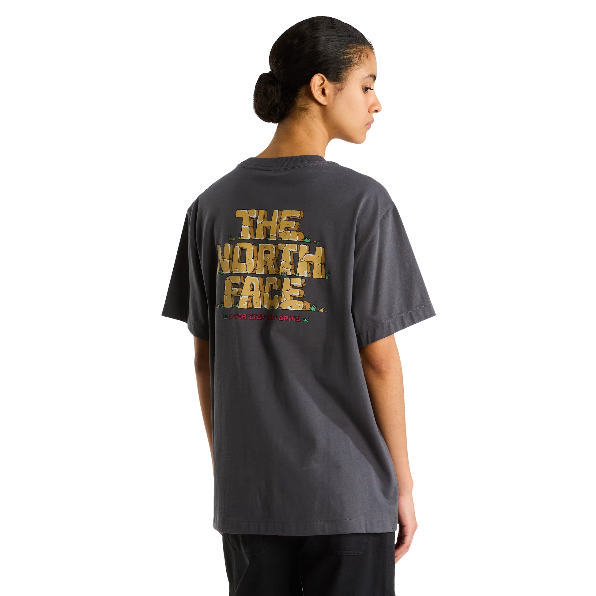 T-Shirt The North Face Stone Logo Relaxed em Anthracite Grey — modelo feminino de costas, cabelo apanhado, calças pretas, gráfico "The North Face" estilo pedra em dourado/ocre e inscrição "Never Stop Exploring" em vermelho nas costas visíveis, fundo branco.