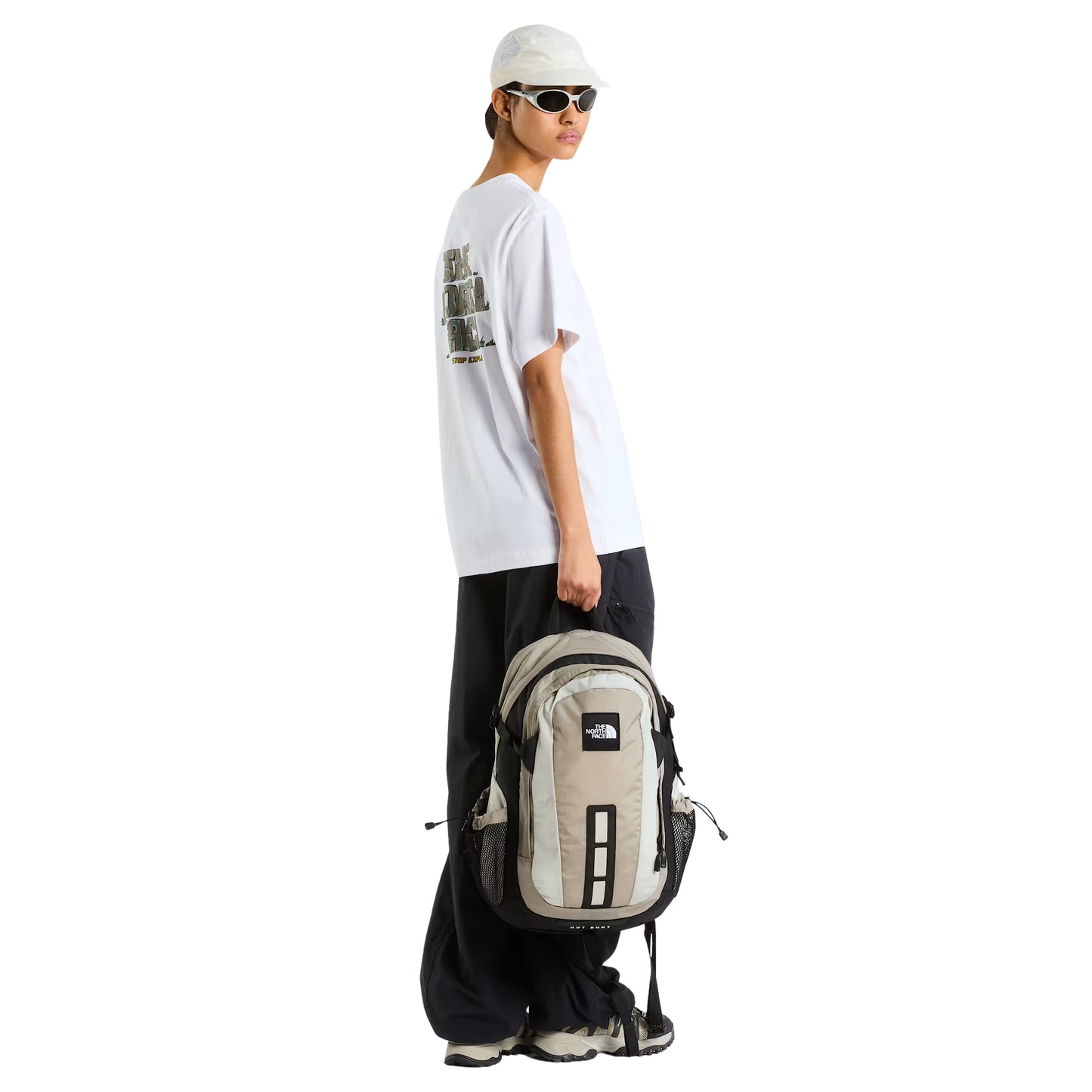 T-Shirt The North Face Stone Logo Relaxed em TNF White — modelo feminino de perfil esquerdo, boné branco TNF e óculos de sol brancos, calças cargo pretas largas e sapatilhas TNF cinza, mochila TNF bege/creme transportada pela alça de mão na mão direita, gráfico Stone Logo parcialmente visível nas costas, fundo branco.