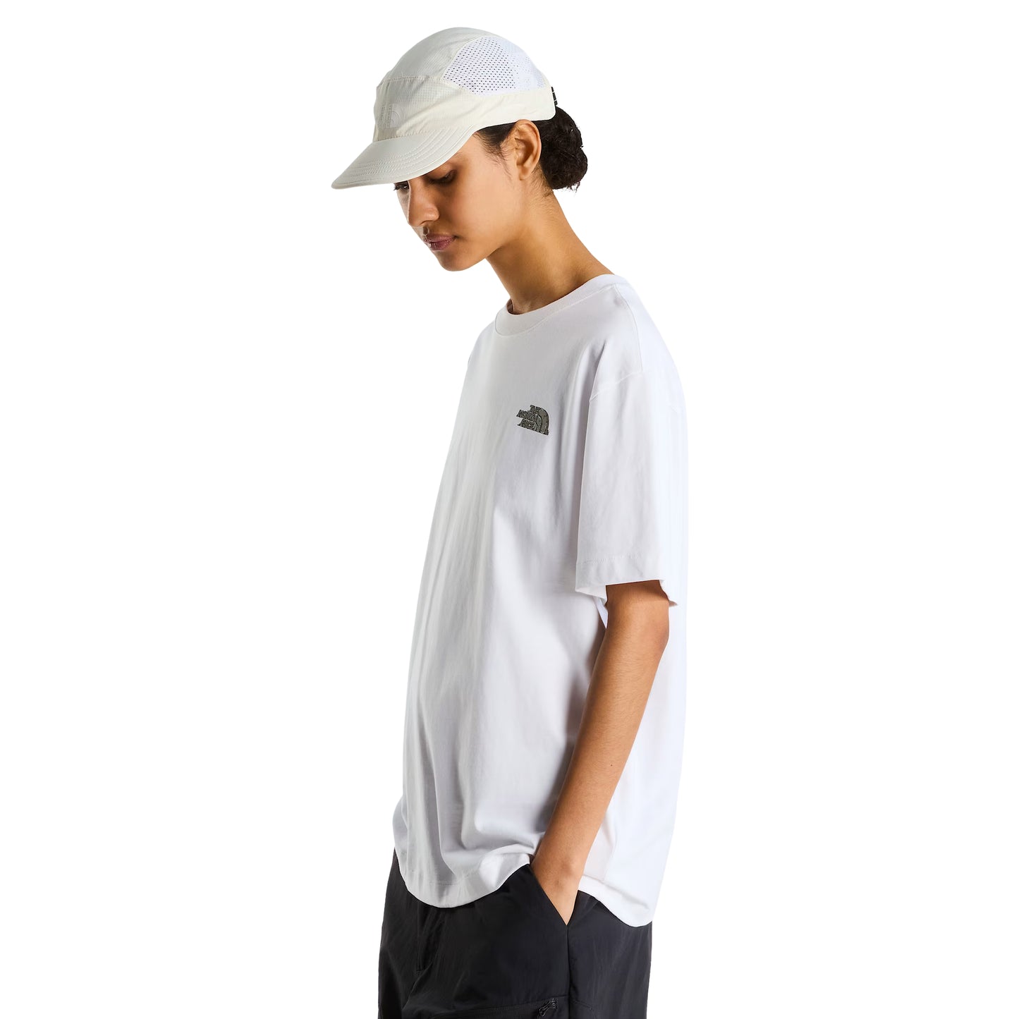 T-Shirt The North Face Stone Logo Relaxed em TNF White — modelo feminino de perfil esquerdo, boné TNF branco perfurado, calças pretas cargo, logo TNF Stone em verde musgo/caqui no peito visível, corte relaxed oversized, fundo branco.