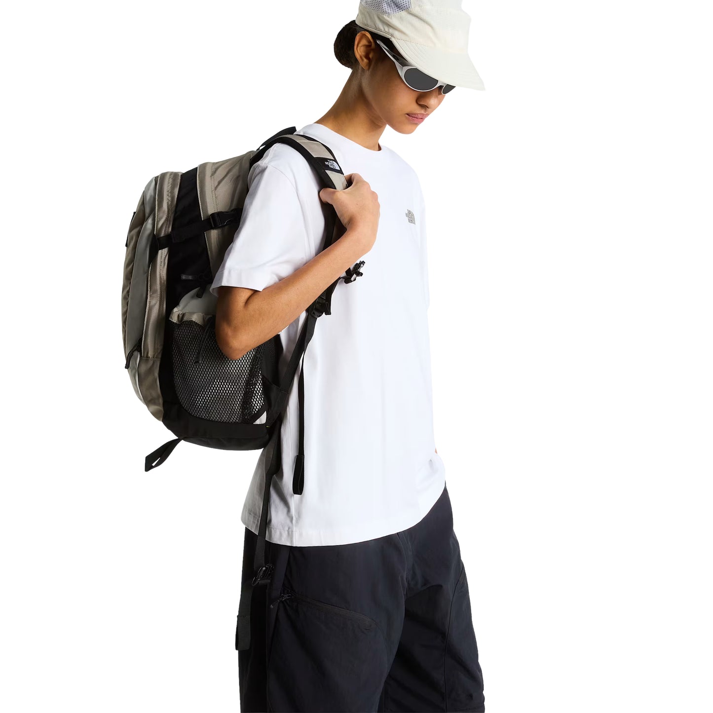 T-Shirt The North Face Stone Logo Relaxed em TNF White — modelo feminino de frente a 3/4, boné branco TNF e óculos de sol brancos, calças cargo pretas largas, mochila TNF cáqui/cinza às costas com bolso lateral em malha preta, logo TNF Stone em verde musgo/cáqui no peito esquerdo visível, fundo branco.