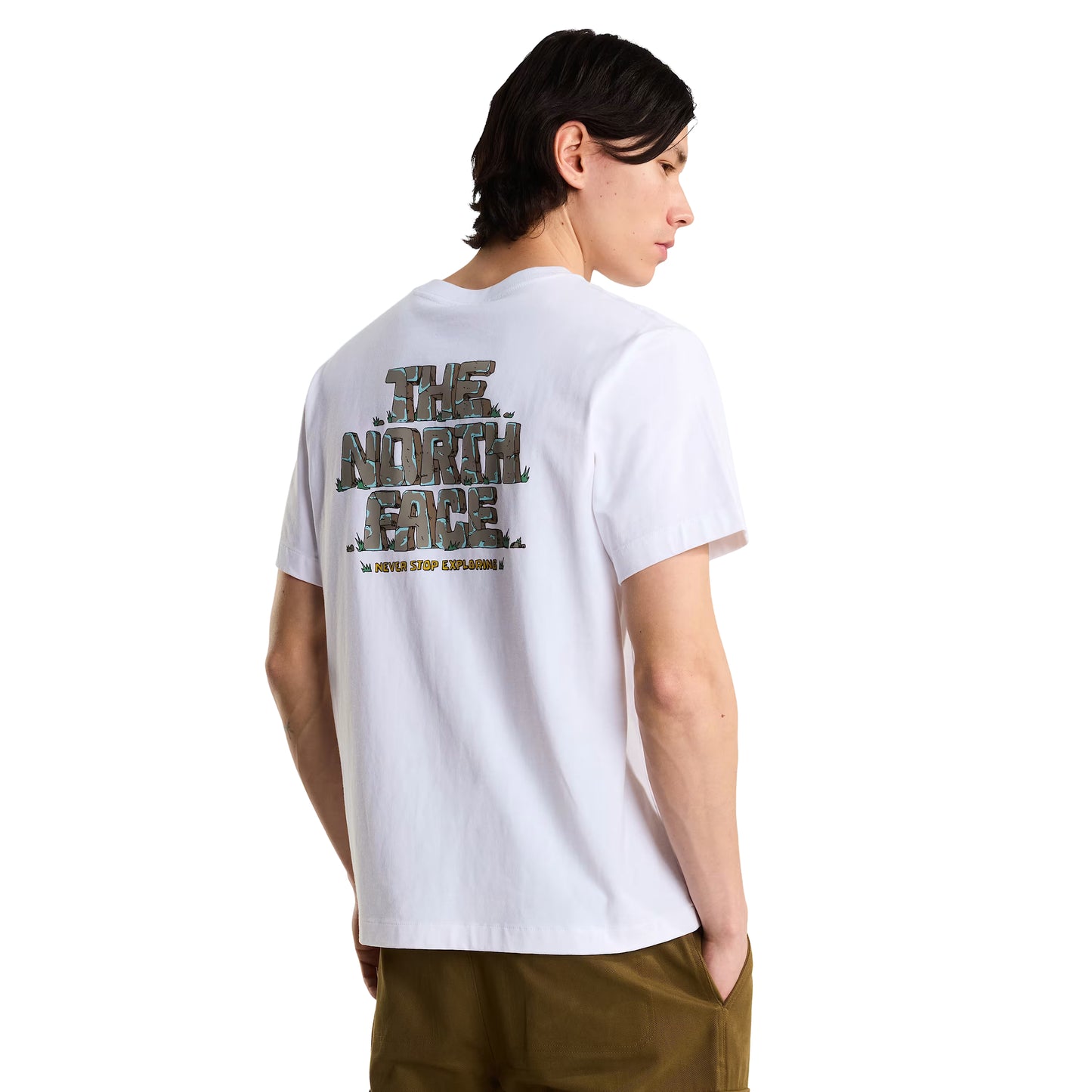T-Shirt The North Face Stone Logo Relaxed em TNF White — modelo masculino de costas a 3/4, cabelo comprido escuro, calças cargo verde-oliva, gráfico "The North Face" estilo pedra em verde musgo/caqui e inscrição "Never Stop Exploring" em amarelo nas costas visíveis, fundo branco.