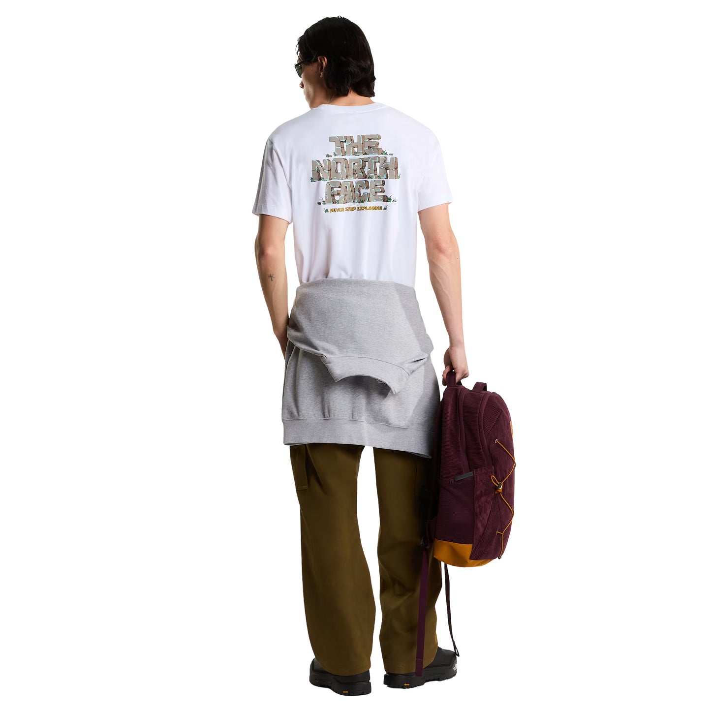 T-Shirt The North Face Stone Logo Relaxed em TNF White — modelo masculino de costas, cabelo comprido escuro e óculos de sol, calças cargo verde-oliva com sweatshirt cinza atada à cintura e mochila TNF bordô/vinho na mão direita, gráfico "The North Face" estilo pedra em dourado/ocre e inscrição "Never Stop Exploring" em amarelo nas costas visíveis, fundo branco.