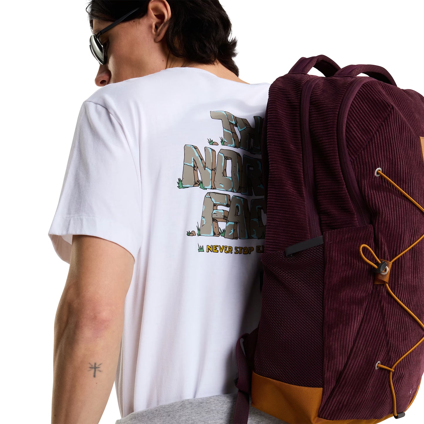 T-Shirt The North Face Stone Logo Relaxed em TNF White — modelo masculino de costas a 3/4, óculos de sol escuros e cabelo comprido escuro, calças cargo verde-oliva, mochila TNF bordô/vinho com detalhes em amarelo-mostarda às costas, gráfico "The North Face" estilo pedra em cinza/verde musgo e inscrição "Never Stop Exploring" em amarelo parcialmente visíveis, fundo branco.