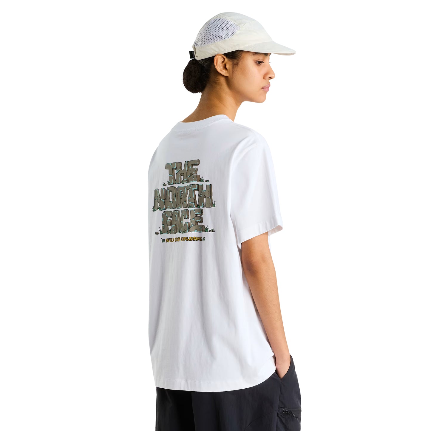 T-Shirt The North Face Stone Logo Relaxed em TNF White — modelo feminino de costas a 3/4, boné TNF branco perfurado, calças pretas cargo, gráfico "The North Face" estilo pedra em verde musgo/caqui e inscrição "Never Stop Exploring" em amarelo nas costas visíveis, fundo branco.