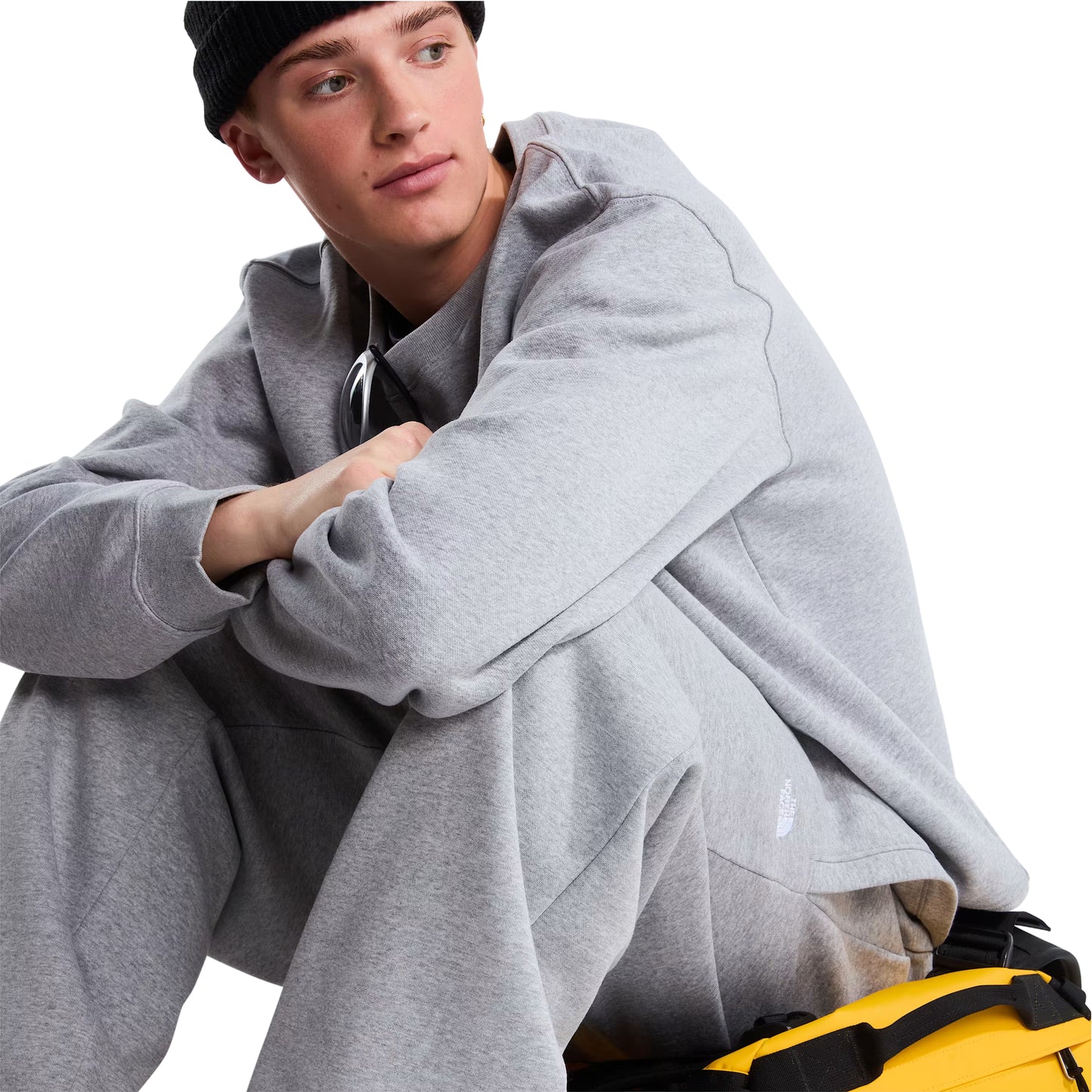 Sweat The North Face TNF Essential Simple Dome Relaxed em TNF Pale Grey Heather — modelo masculino sentado de frente a 3/4, gorro preto TNF, mochila duffel TNF amarela e preta como assento, corte relaxado com volume generoso nas mangas e logótipo TNF half-dome bordado em branco visível, fundo branco.