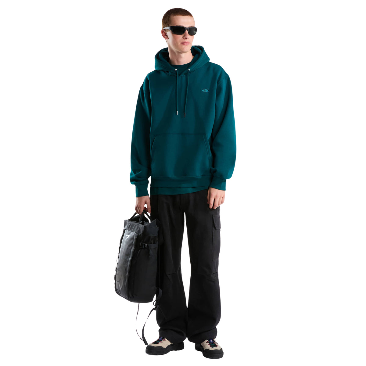 Modelo de corpo inteiro com sweat The North Face Essential Simple Dome Pacific Teal, calças pretas e mochila na mão.