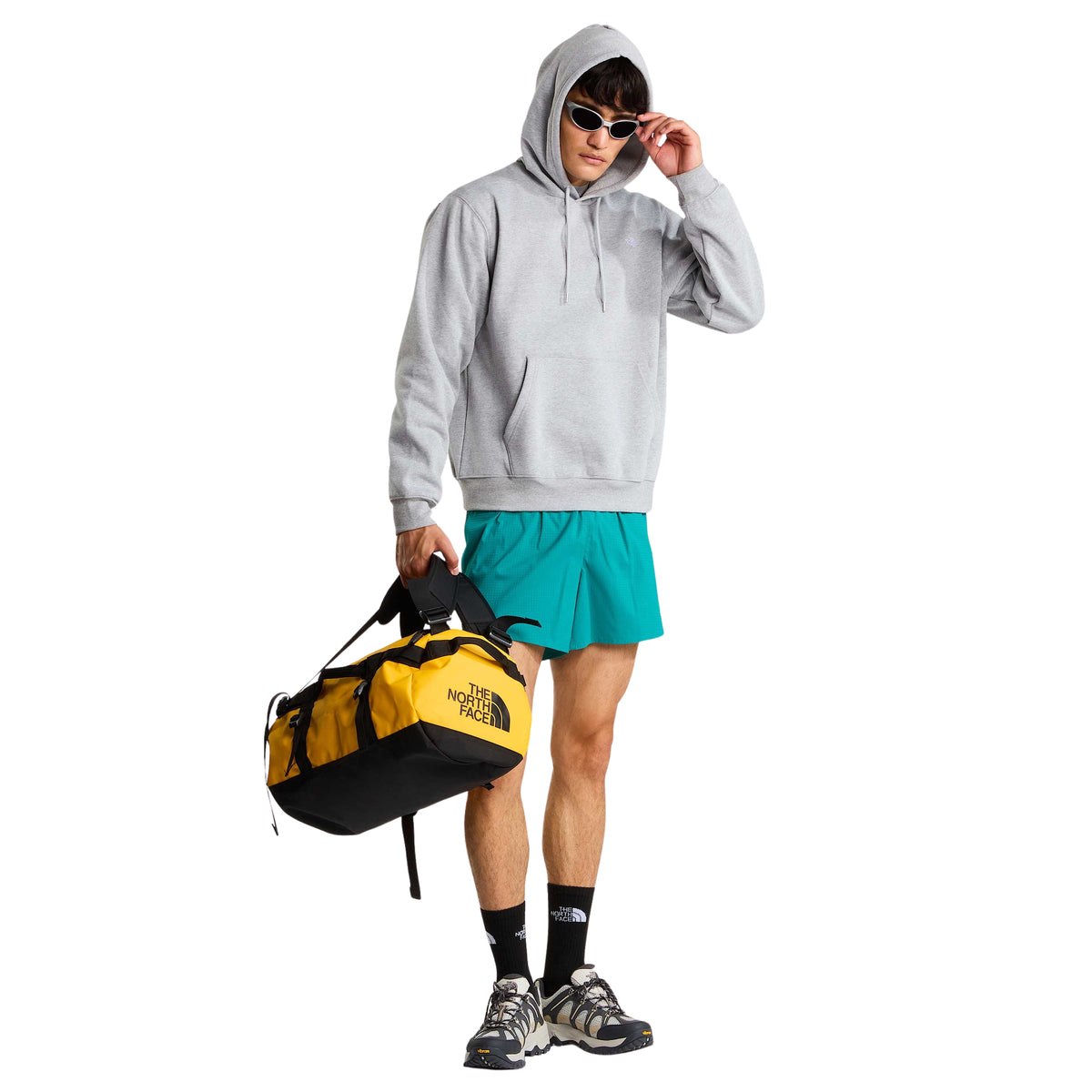 Modelo de corpo inteiro com sweat The North Face Essential Simple Dome cinza-claro mesclado, capuz colocado, calções turquesa e saco amarelo.