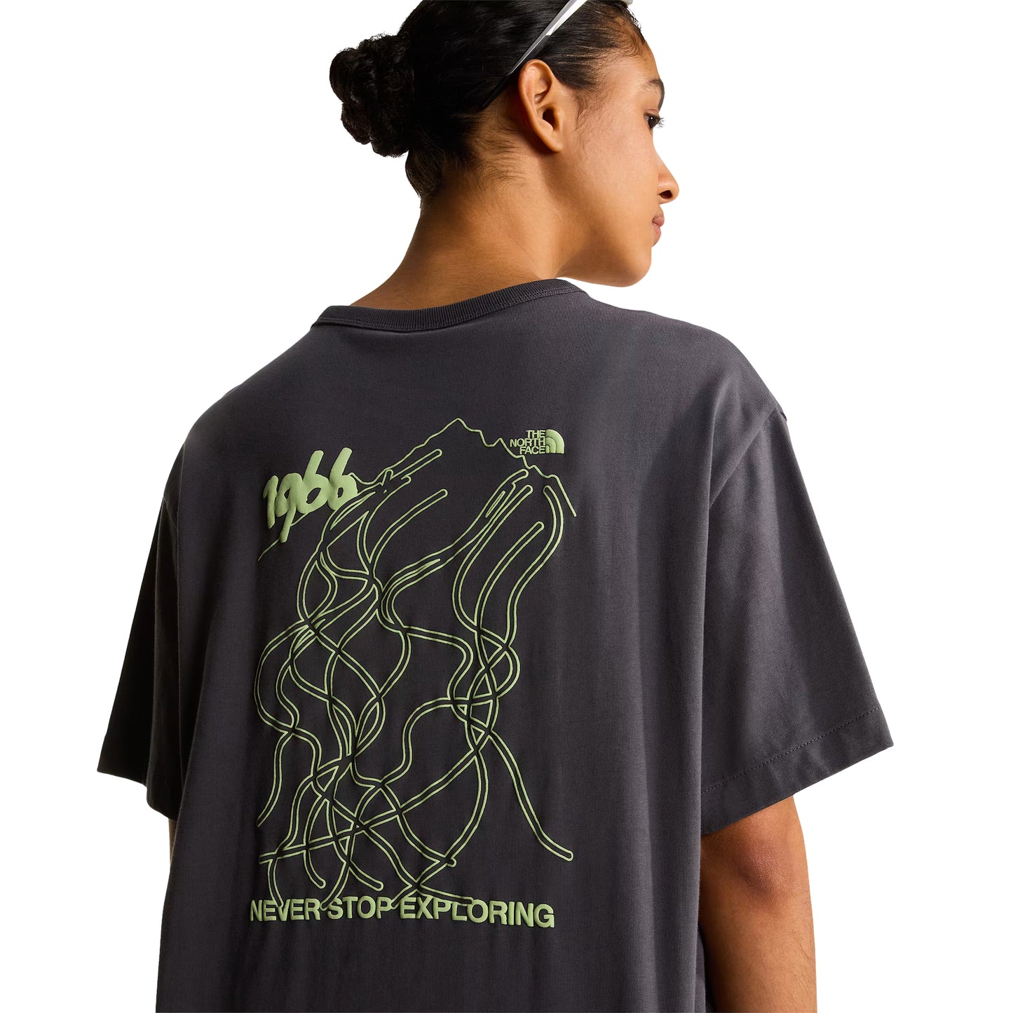 T-shirt unissexo The North Face NSE Slopes Relaxed em Anthracite Grey — modelo feminino de costas a 3/4, headband branca, cabelo apanhado, detalhe do gráfico NSE Slopes "1966 Never Stop Exploring" estampado em verde lima nas costas, logótipo TNF half-dome no canto superior direito do gráfico, fundo branco.