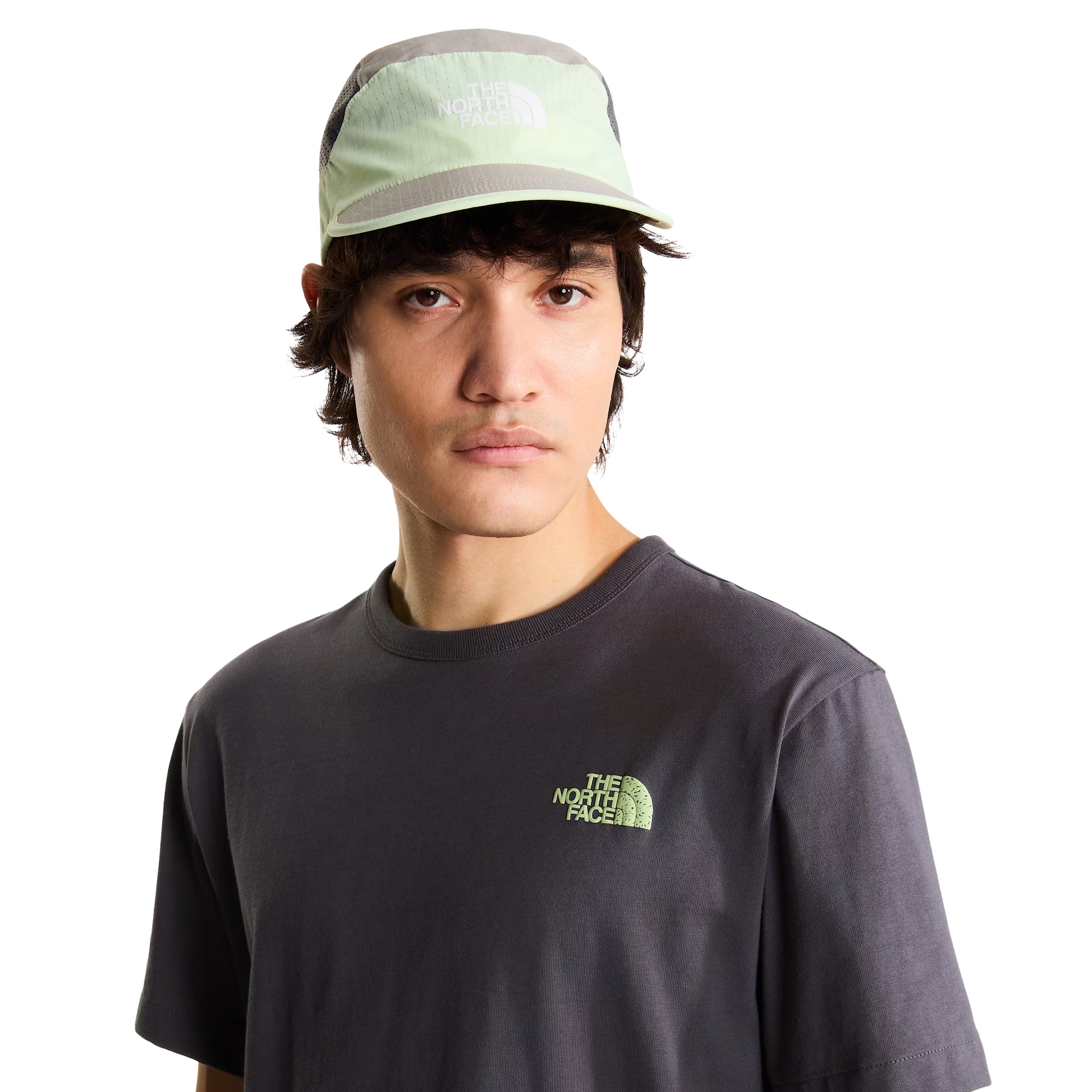 T-shirt unissexo The North Face NSE Slopes Relaxed em Anthracite Grey — modelo masculino de frente a 3/4, boné técnico branco e cinza TNF, calças cargo verde-oliva, logótipo "The North Face" half-dome estampado em relevo verde lima no peito esquerdo, gola canelada e jersey Anthracite Grey visíveis, fundo branco.