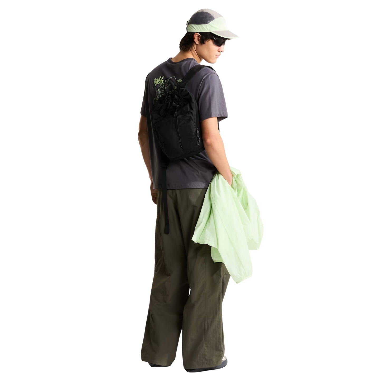 -shirt unissexo The North Face NSE Slopes Relaxed em Anthracite Grey — modelo masculino de costas, corpo inteiro, boné técnico branco e cinza TNF, calças cargo verde-oliva, gráfico NSE Slopes "1966 Never Stop Exploring" estampado em verde lima nas costas, fundo branco.