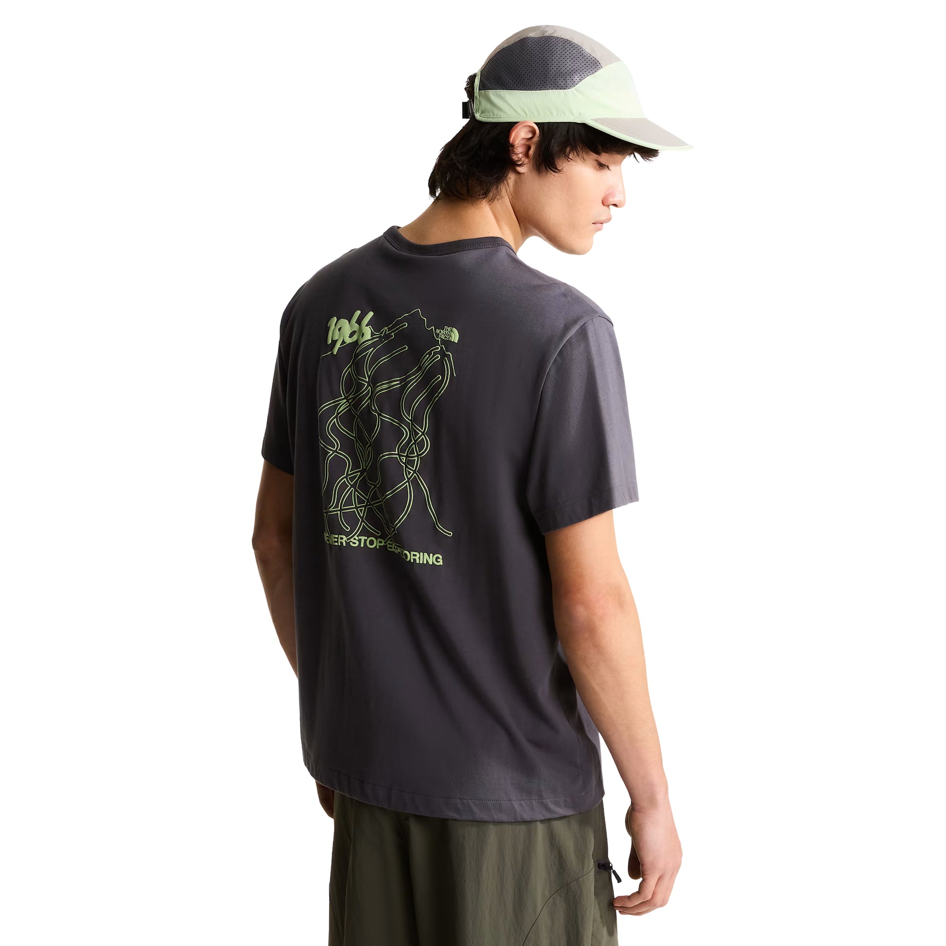 T-shirt unissexo The North Face NSE Slopes Relaxed em Anthracite Grey — modelo masculino de costas a 3/4, boné técnico branco e cinza TNF, calças cargo verde-oliva, gráfico NSE Slopes "1966 Never Stop Exploring" estampado em verde lima nas costas, fundo branco.
