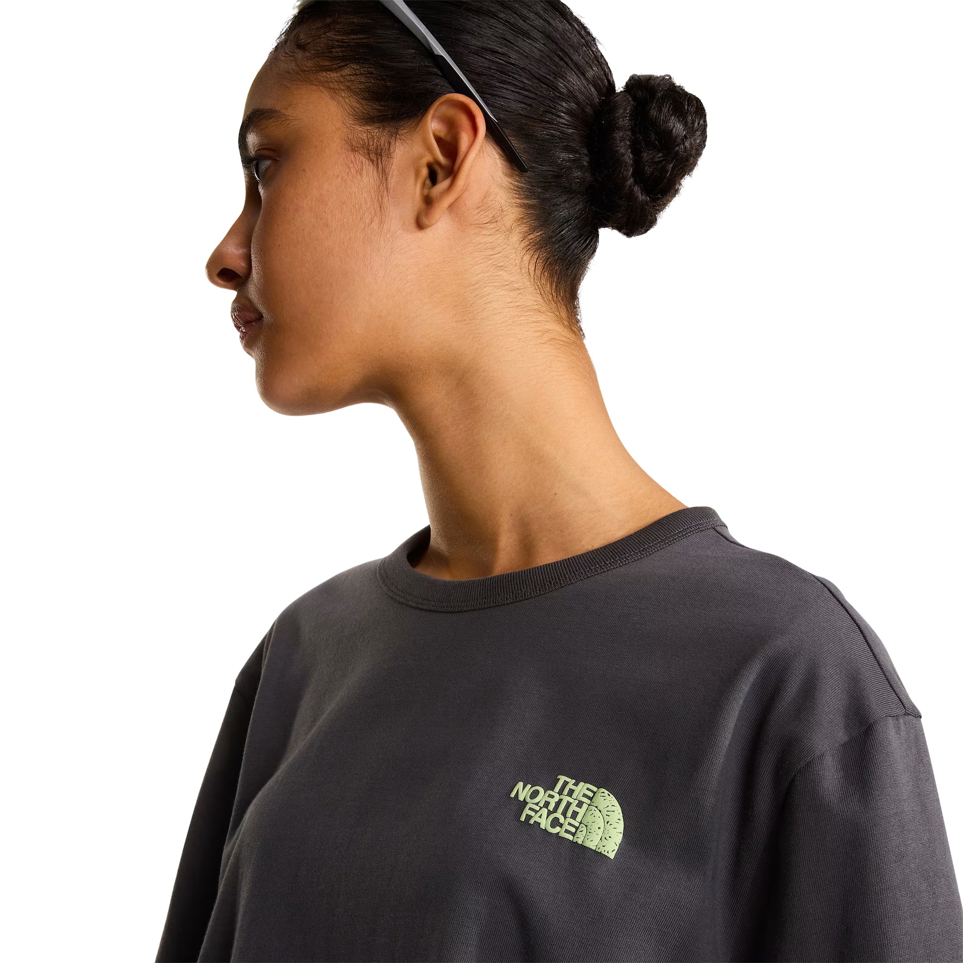 T-shirt unissexo The North Face NSE Slopes Relaxed em Anthracite Grey — modelo feminino de frente, meio corpo, óculos de sol brancos na cabeça, calças cargo verde-oliva, logótipo "The North Face" half-dome estampado em relevo verde lima no peito esquerdo, gola canelada e jersey Anthracite Grey visíveis, fundo branco.