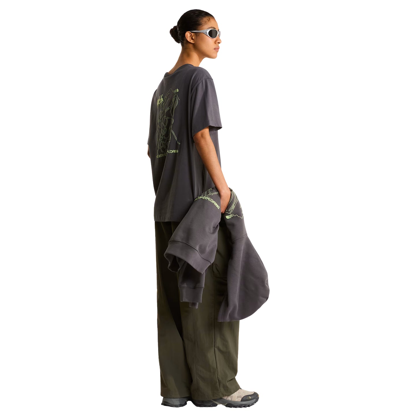 T-shirt unissexo The North Face NSE Slopes Relaxed em Anthracite Grey — modelo feminino de costas a 3/4, óculos de sol brancos na cabeça, calças cargo verde-oliva, gráfico NSE Slopes "1966 Never Stop Exploring" estampado em verde lima nas costas, corte relaxado com ombros descaídos visíveis, fundo branco.