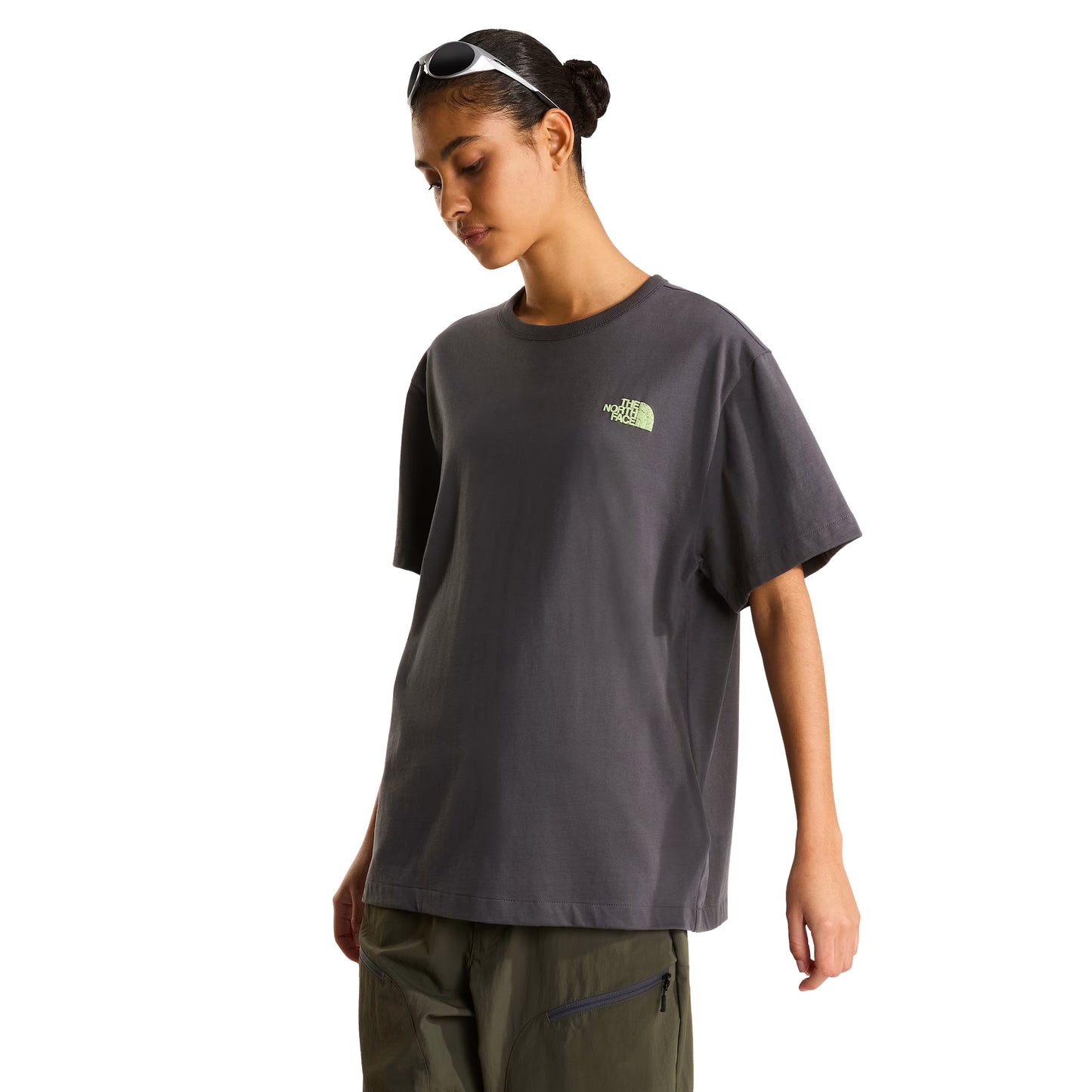 T-shirt unissexo The North Face NSE Slopes Relaxed em Anthracite Grey — modelo feminino de frente a 3/4, óculos de sol brancos na cabeça, calças cargo verde-oliva, logótipo "The North Face" half-dome estampado em relevo verde lima no peito esquerdo, corte relaxado com ombros descaídos visíveis, fundo branco.