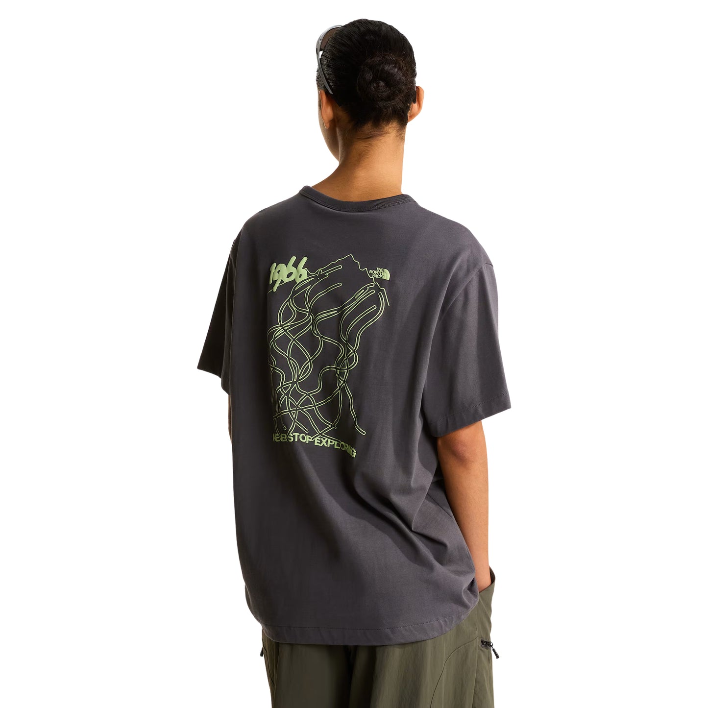 T-shirt unissexo The North Face NSE Slopes Relaxed em Anthracite Grey — modelo feminino de costas, boné preto TNF, cabelo apanhado, calças cargo verde-oliva, gráfico NSE Slopes "1966 Never Stop Exploring" estampado em verde lima nas costas, corte relaxado com ombros descaídos visíveis, fundo branco.