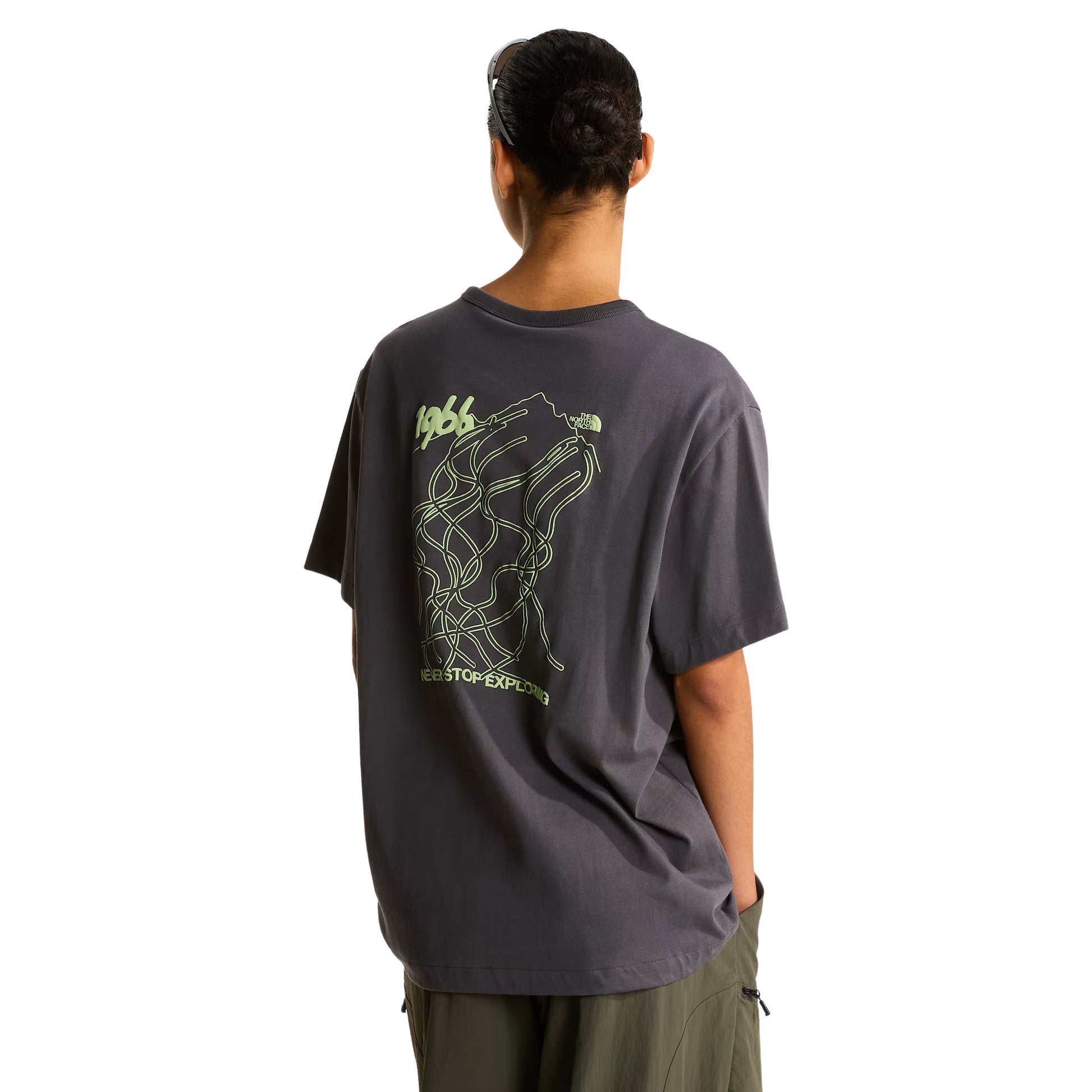 T-shirt unissexo The North Face NSE Slopes Relaxed em Anthracite Grey — modelo feminino de costas, boné preto TNF, cabelo apanhado, calças cargo verde-oliva, gráfico NSE Slopes "1966 Never Stop Exploring" estampado em verde lima nas costas, corte relaxado com ombros descaídos visíveis, fundo branco.