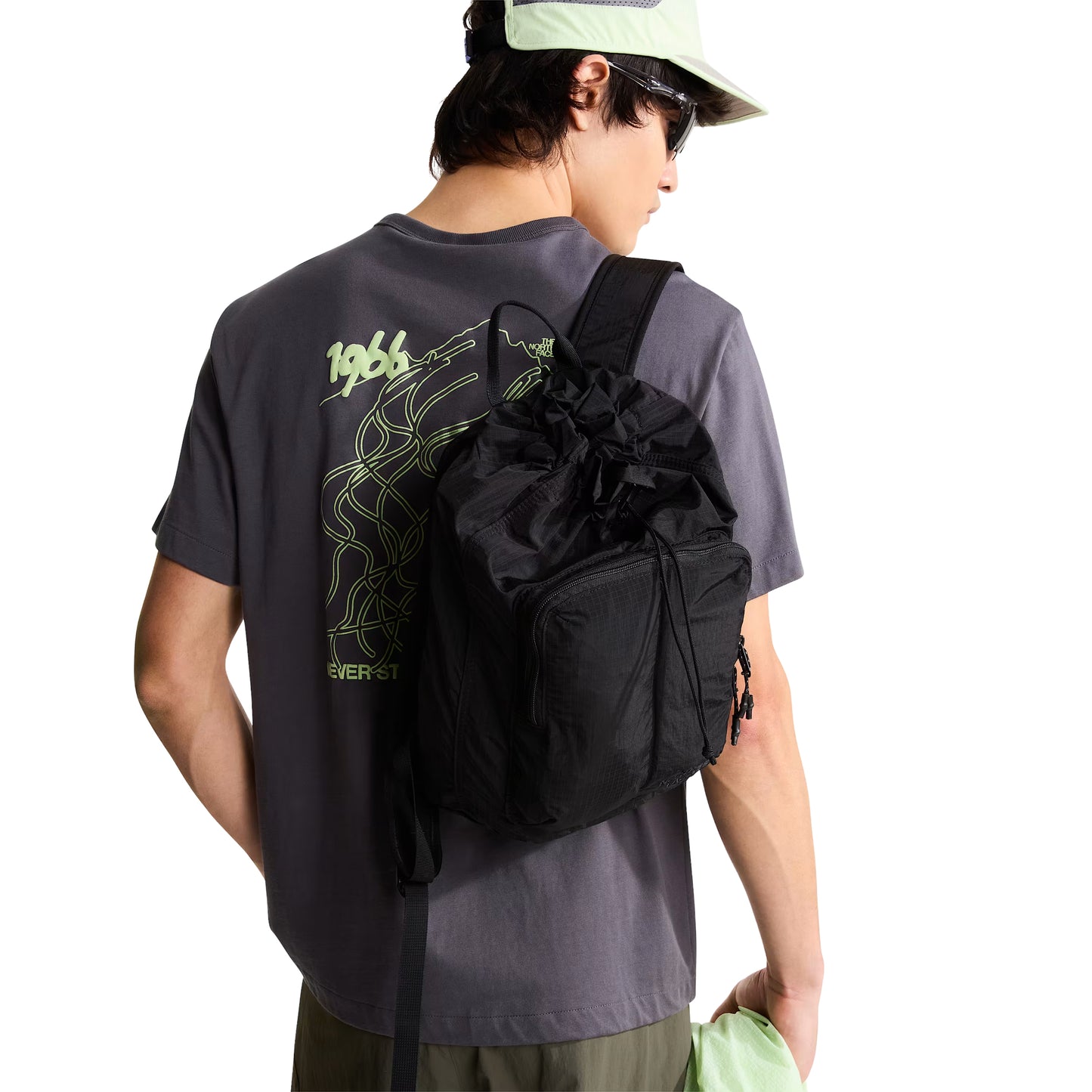 T-shirt unissexo The North Face NSE Slopes Relaxed em Anthracite Grey — modelo masculino de costas, meio corpo, boné técnico verde-água TNF, óculos de sol, mochila sling TNF preta ao ombro, calções cargo verde-oliva, gráfico NSE Slopes "1966 Never Stop Exploring" estampado em verde lima nas costas parcialmente visível, fundo branco.