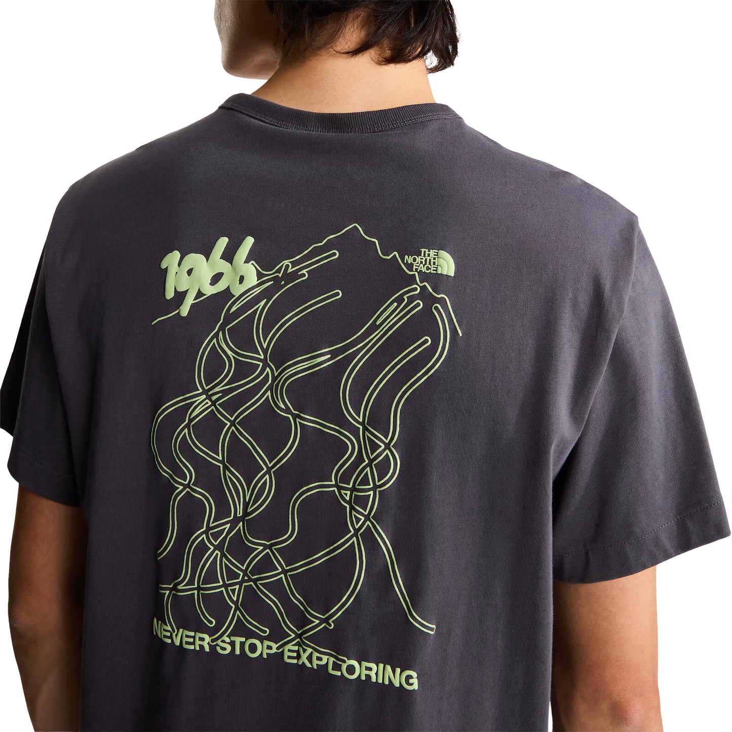 T-shirt unissexo The North Face NSE Slopes Relaxed em Anthracite Grey — modelo masculino de costas, meio corpo, detalhe do gráfico NSE Slopes "1966 Never Stop Exploring" estampado em verde lima nas costas, ilustração de pistas de ski entrelaçadas e logótipo TNF half-dome no canto superior direito visíveis, fundo branco.
