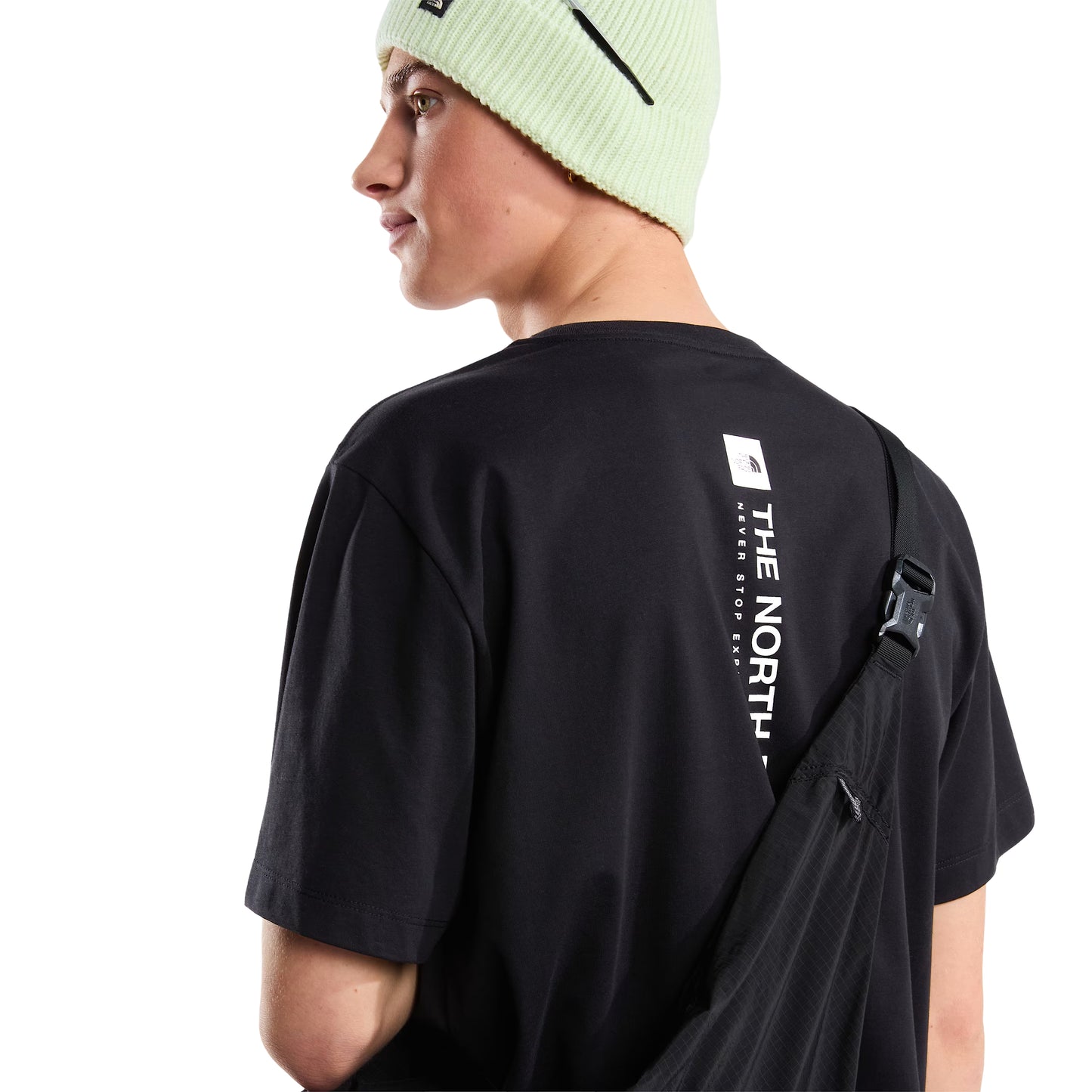 T-Shirt The North Face Vertical Regular em TNF Black — modelo masculino de costas a 3/4, gorro TNF verde-lima, mochila preta de tiracolo com fivela preta, gráfico vertical "The North Face" em branco parcialmente visível nas costas, fundo branco.