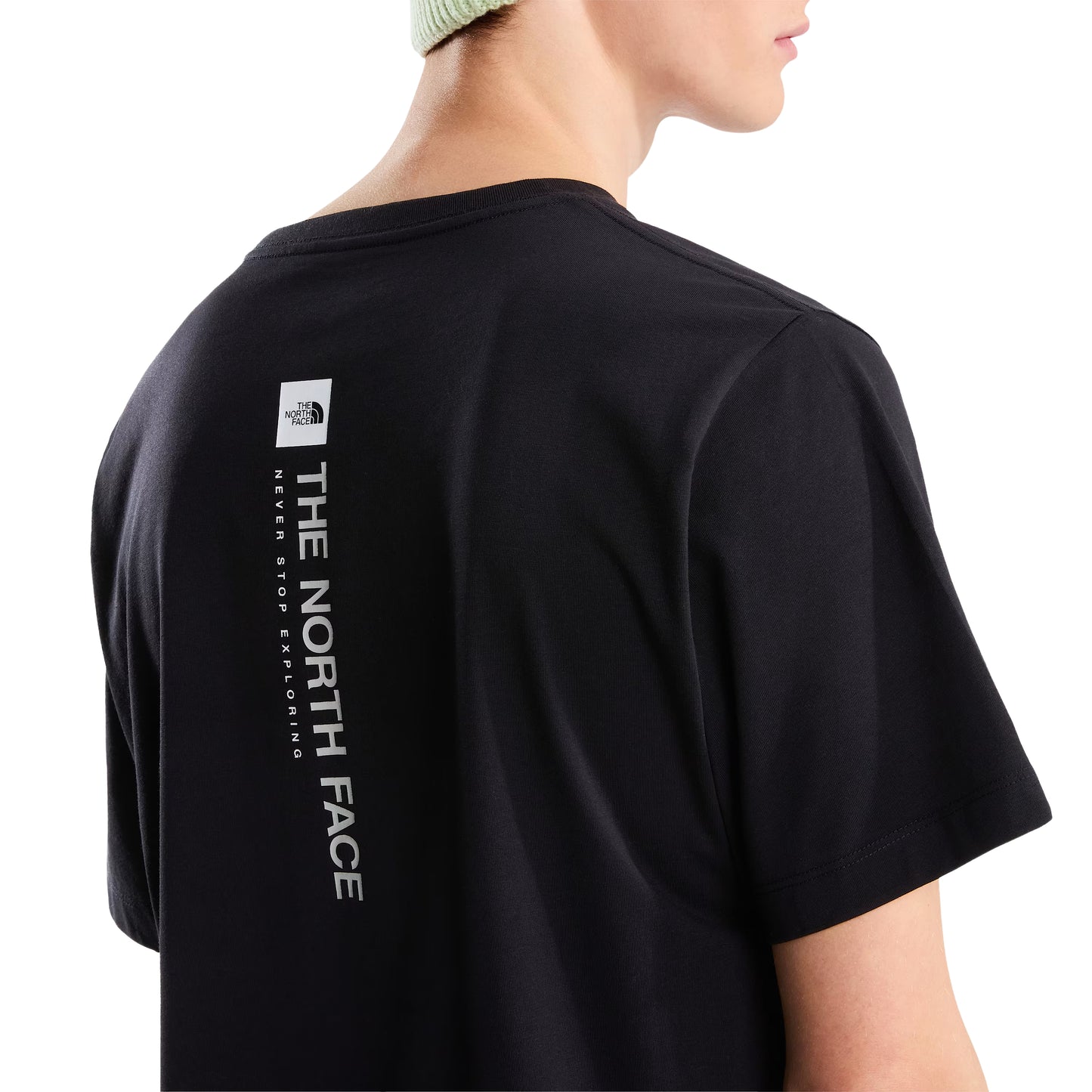 T-Shirt The North Face Vertical Regular em TNF Black — detalhe das costas de modelo masculino, gráfico vertical "The North Face" em branco com logo TNF em caixa branca no topo e inscrição "Never Stop Exploring" visíveis em close-up, gorro verde-lima, fundo branco.