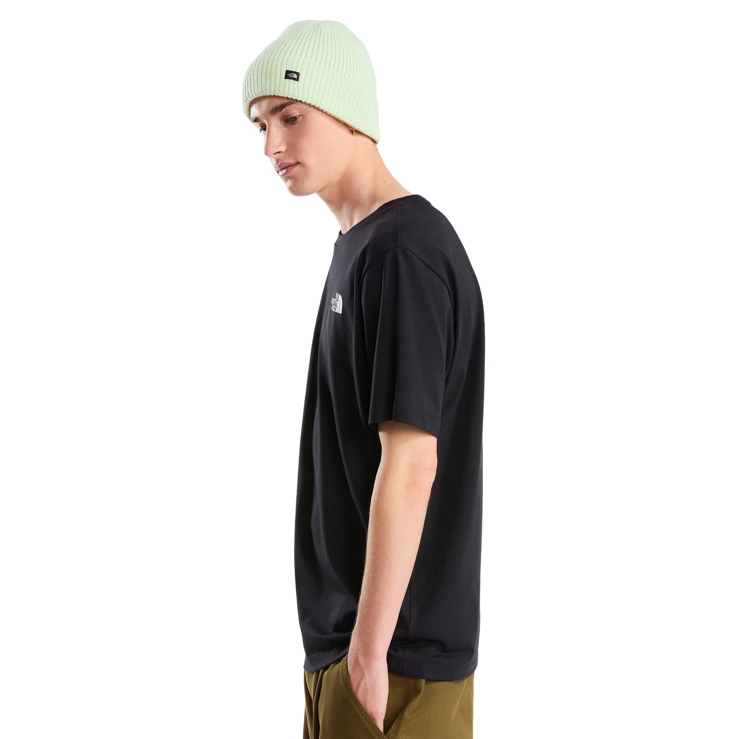 T-Shirt The North Face Vertical Regular em TNF Black — modelo masculino de frente a 3/4, gorro TNF verde-lima, calças cargo bege/cáqui largas, logo TNF half-dome em branco no peito esquerdo visível, corte regular, fundo branco.