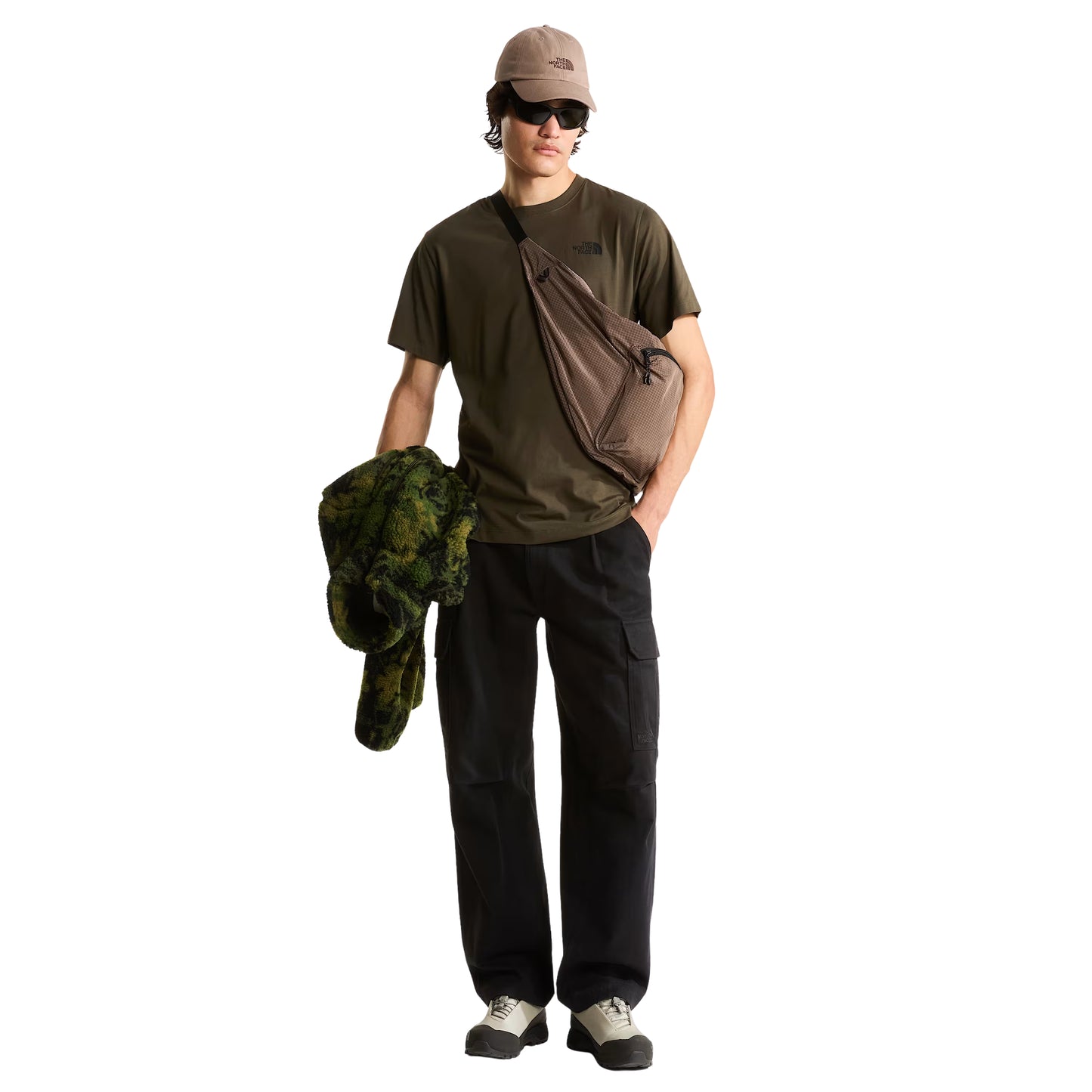 T-Shirt The North Face Yosemite Oak Box NSE Regular em New Taupe Green — modelo masculino de frente, corpo inteiro, boné TNF bege/taupe, óculos de sol escuros, mochila sling TNF bege/taupe em tiracolo, calças cargo pretas largas e sapatilhas cinza-claro, casaco fleece camuflado verde na mão esquerda, fundo branco.