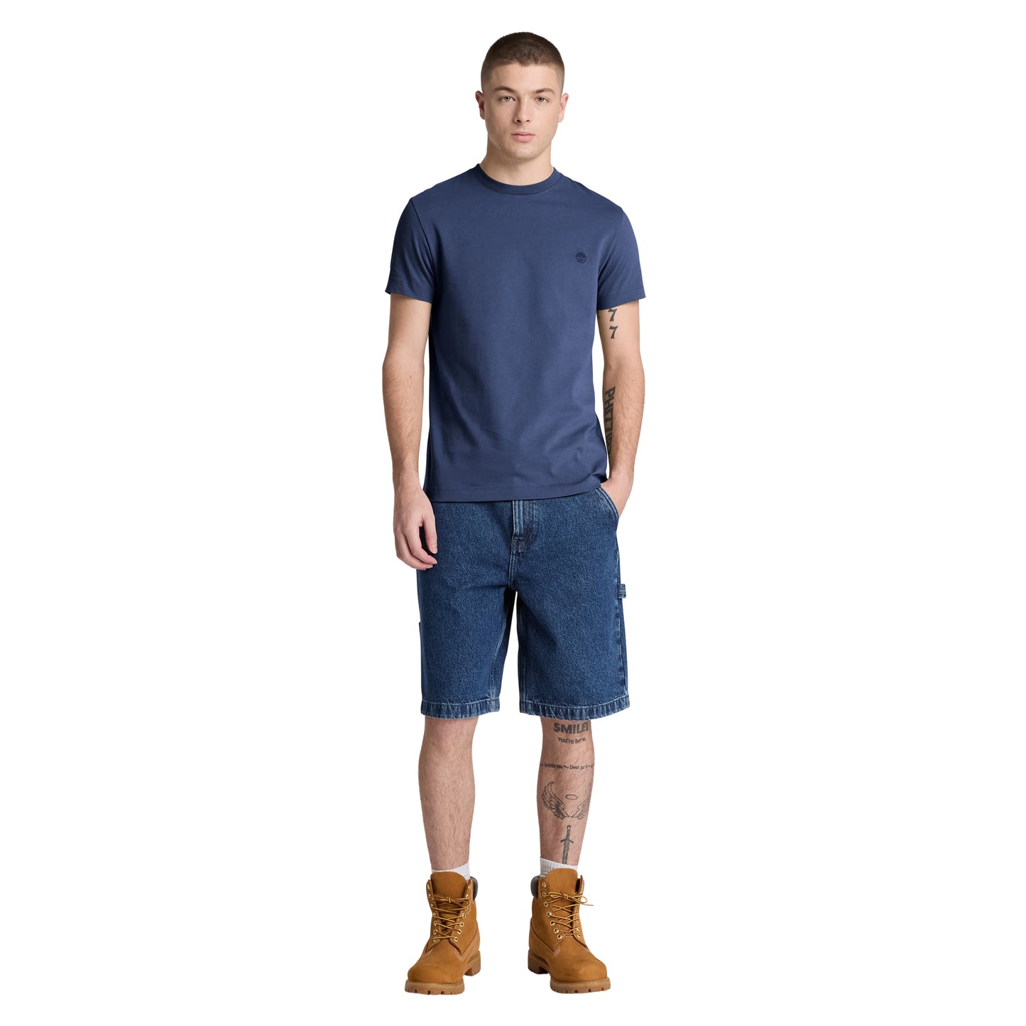 T-shirt Timberland Dunstan River em Naval Academy — vista frontal com modelo, corpo inteiro, corte slim e comprimento ligeiramente curto visíveis, combinado com calções denim azul e botas wheat Timberland.