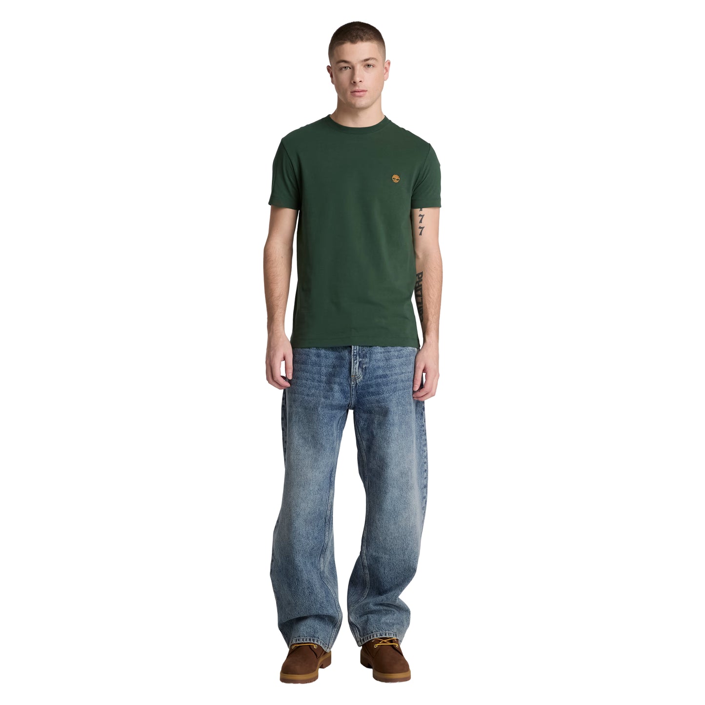 T-shirt Timberland Dunstan River em Sycamore — vista frontal com modelo, corpo inteiro, corte slim e comprimento ligeiramente curto visíveis, combinado com calças de ganga azul largas e botas wheat Timberland.