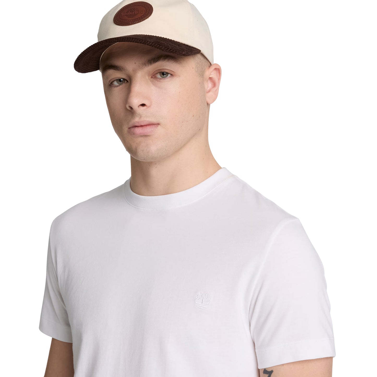 T-shirt Timberland Dunstan River em White — detalhe do logótipo árvore Timberland bordado em branco tonal no peito esquerdo e gola redonda canelada, close-up com modelo com boné Timberland ecru/castanho.